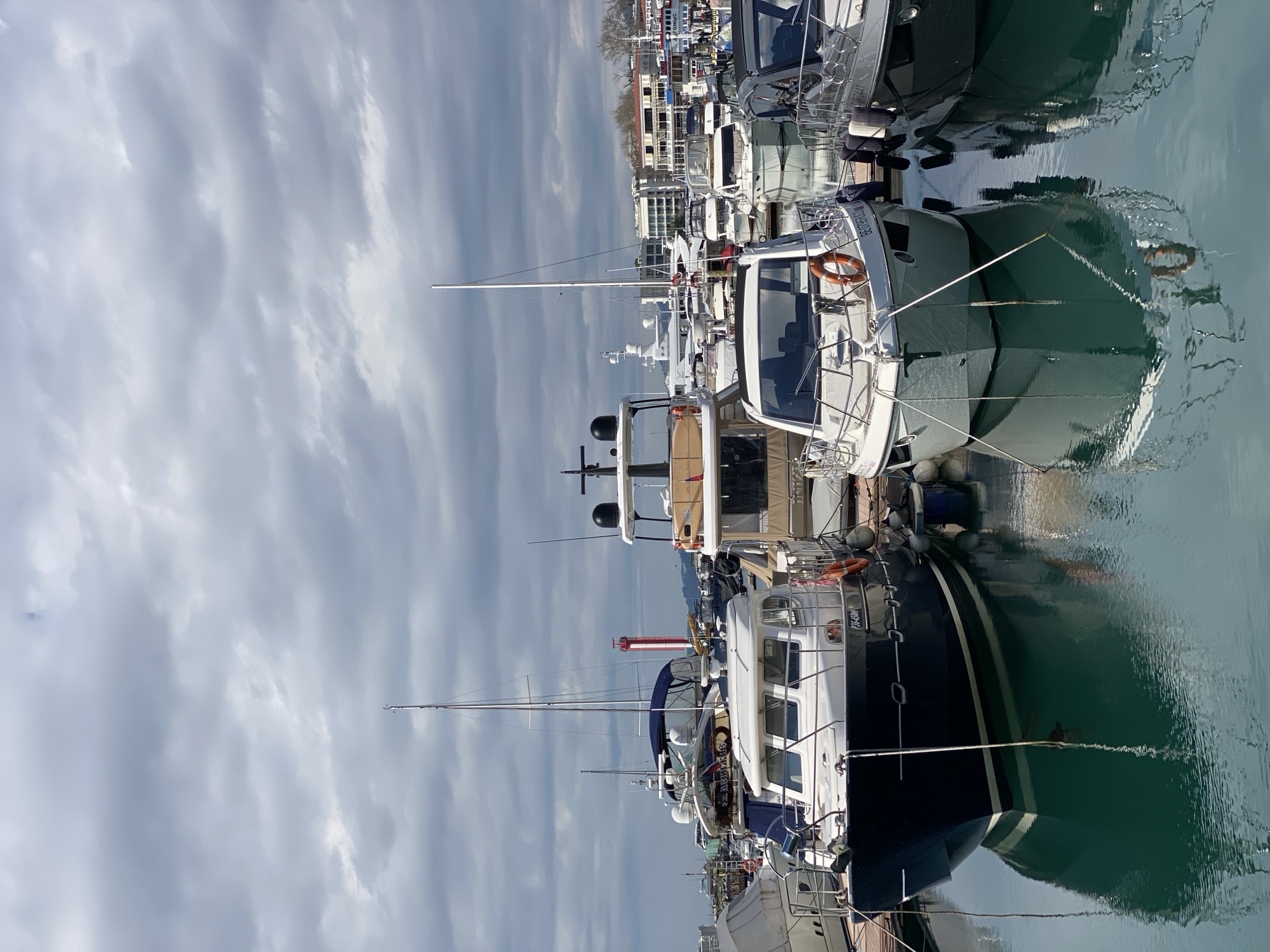 The image captures a marina in Sochi, Russia, featuring a dense collection of boats moored alongside each other. The foreground is dominated by several yachts and motorboats, varying in size and design, their hulls reflecting in the calm water. Masts of the boats extend upwards, puncturing a cloudy sky, the sky suggesting an overcast day. In the background, a row of buildings lines the waterfront, indicative of the urban landscape of Sochi. The buildings appear to be a mix of residential and commercial structures. The overall scene presents a typical marina setting within a city.