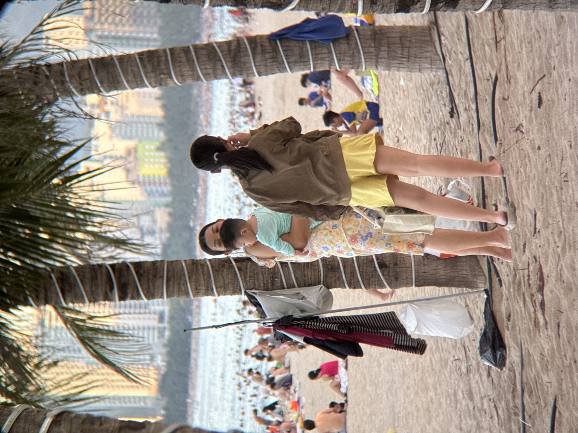This is a medium shot, taken from a low angle, of a crowded beach in Nha Trang, Vietnam. The foreground is dominated by the sandy beach and the trunks of two palm trees, wrapped with white rope. A woman in a floral dress is holding a baby wearing a light blue shirt. To her right, a young woman wearing a brown jacket and yellow shorts is standing, facing away from the camera, with her hand to her ear. Behind them, the ocean is visible, filled with many people swimming and sunbathing. In the background, tall buildings line the coast, suggesting an urban setting. There are various objects scattered on the sand, including clothing hanging on a pole, a white plastic bag, and a black garbage bag. The scene depicts a typical day at a bustling tourist beach.