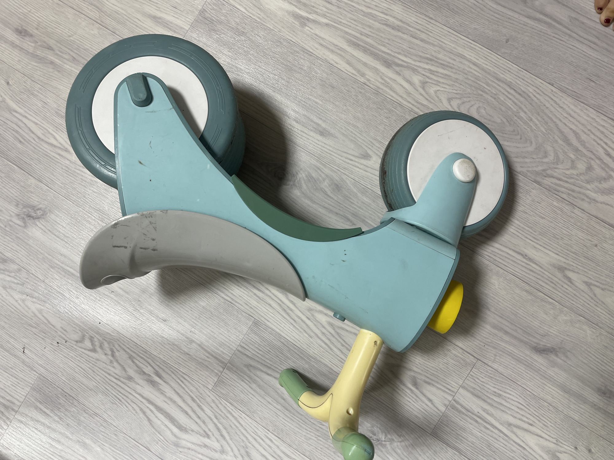 An overhead shot shows a light blue balance bike on a gray wood-grain floor. The bike has two wheels, one larger than the other, with white inner circles and light gray tires. The frame is light blue, with a gray section at the front and a dark green section where the seat would be. The handlebars are yellow and light green. There is no indication of the location being Baranovka, Russia. No people are visible in the immediate vicinity of the bike, though a foot is visible in the upper right.