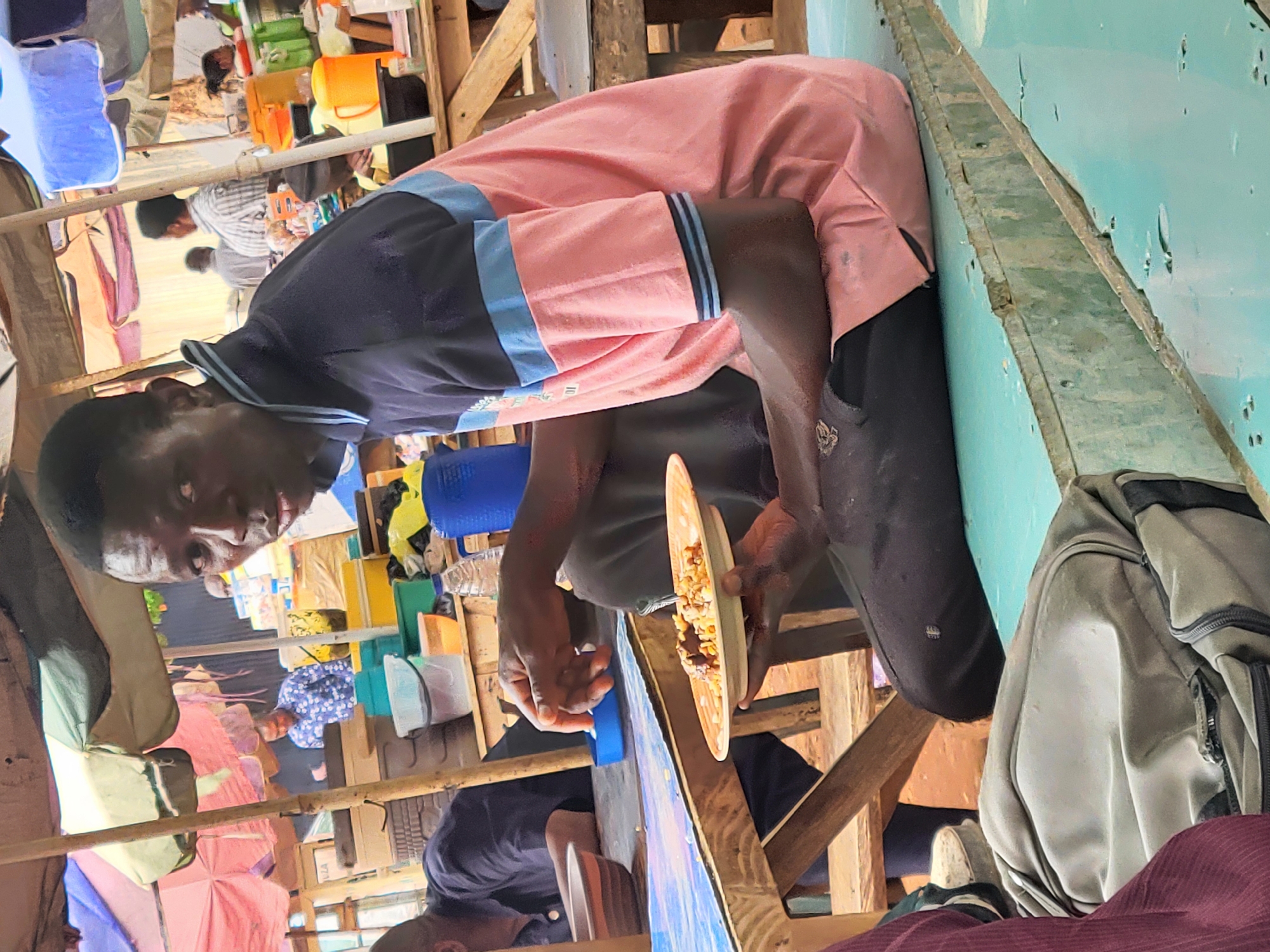 In an indoor setting that appears to be a market or makeshift eatery in Abuja, Nigeria, a man is the primary subject. He holds a plate of food, seemingly engaged in eating. He wears a two-toned shirt, light pink on top and dark blue on the shoulders, along with dark pants. The surrounding area is cluttered with various items, including plastic containers, bags, and wooden structures. Other people are faintly visible in the background, suggesting a busy public place. The environment looks somewhat improvised, with the eatery constructed from basic materials.