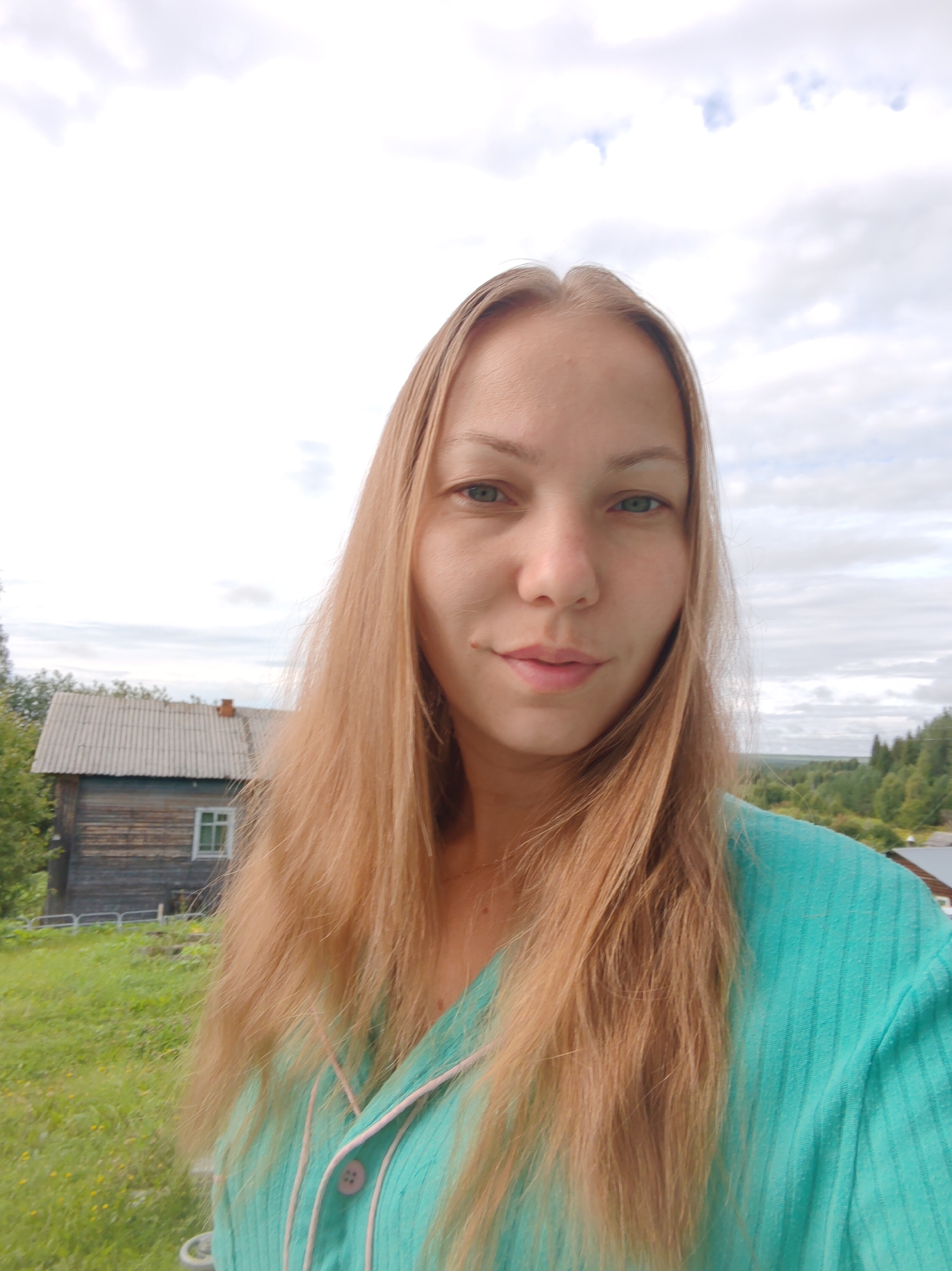 A woman with long blonde hair is pictured outdoors, facing the camera. She is wearing a turquoise shirt with pink trim and buttons. The background shows a wooden house with a grey roof, a green grassy hill, and trees. The sky is partly cloudy. The location appears to be rural.