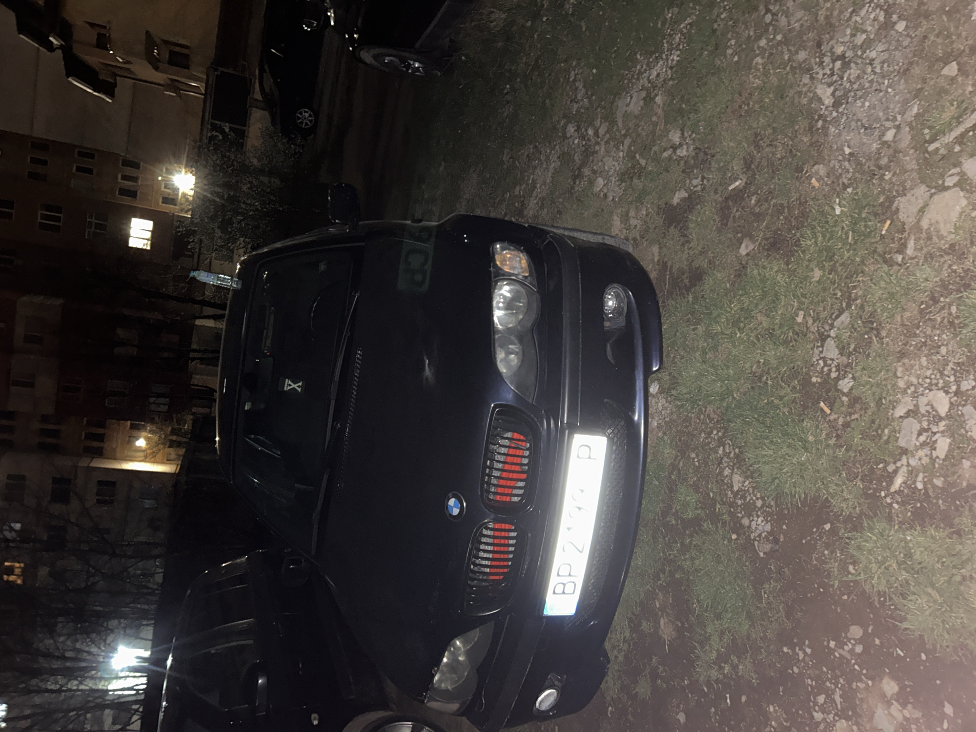 The image depicts a dark blue BMW parked on a patch of ground covered in grass and gravel. The front of the car is facing the viewer, with the license plate visible. The license plate reads "BP 2739 P," indicating it is registered in Sofia, Bulgaria. The background shows an apartment building, illuminated by streetlights and some windows, suggesting the scene is in an urban environment. Given the license plate information, it can be said it is in an urban environment in Bulgaria. Without further information, the location in the city of Vratsa, Bulgaria cannot be confirmed.