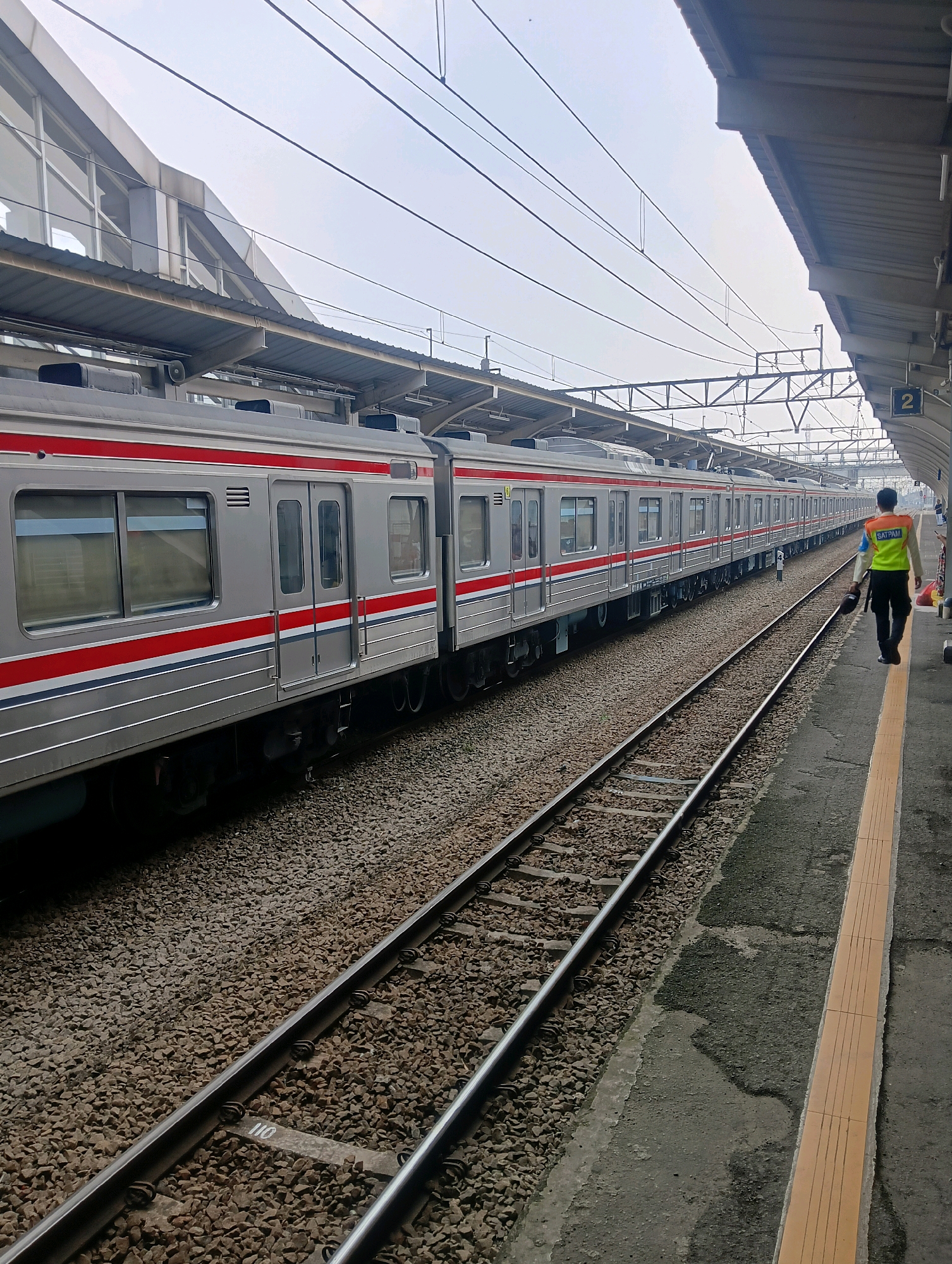 The image depicts a train at the Parung Panjang train station in Indonesia. A commuter train, silver with red and blue stripes, is stopped at the platform. The train cars have large windows and multiple doors. A railway worker wearing a high-visibility vest over a light-colored shirt and dark pants is walking along the platform, carrying a dark cap. The platform has a yellow tactile paving strip. Train tracks extend into the distance, flanked by ballast. Overhead power lines and station infrastructure are visible in the background under an overcast sky.