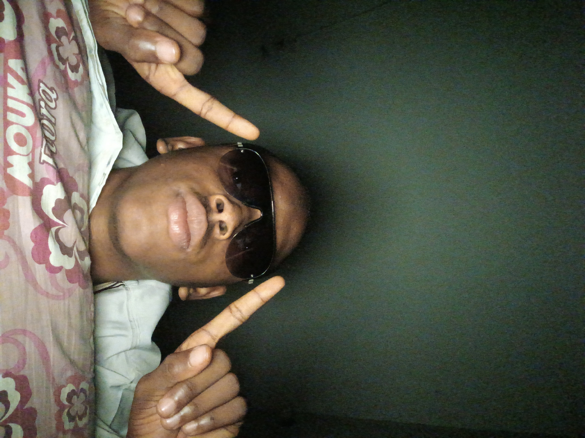 A low-angle, medium shot captures a Black male from the chest up, posing for a selfie. He is wearing dark sunglasses and a light-colored hooded sweatshirt. His left hand is raised, with his index finger pointing towards the camera, and his right hand is also raised, with his index finger pointing upwards. The background appears to be a dark, textured wall or surface. In the foreground, a floral-patterned fabric, possibly a bedsheet, is visible. The lighting is dim, with the subject's face illuminated from the front. The image does not contain any specific details that would allow for the determination of the location in Lagos, Nigeria.