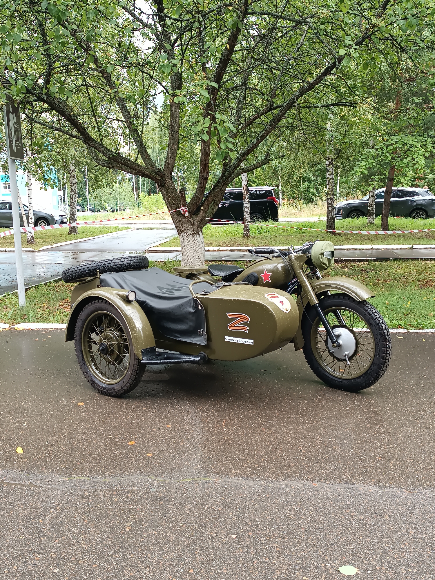 An olive-drab, vintage Soviet-era motorcycle with a sidecar is parked on a wet asphalt road under a leafy tree. The motorcycle features a spare tire mounted on the back of the sidecar. A red star emblem is visible on the motorcycle's fuel tank, and a white "Z" symbol with text below it adorns the sidecar. In the background, several modern cars are parked along a sidewalk. The scene appears to be in an urban or suburban area, possibly a park or residential street, given the presence of trees, sidewalk, and parked vehicles. The overall condition of the motorcycle suggests it might be a preserved or restored vehicle, possibly part of a display or event. The wet road indicates recent rain. There are no people visible in the image. The location is identified as Chaikovsky, Russia.