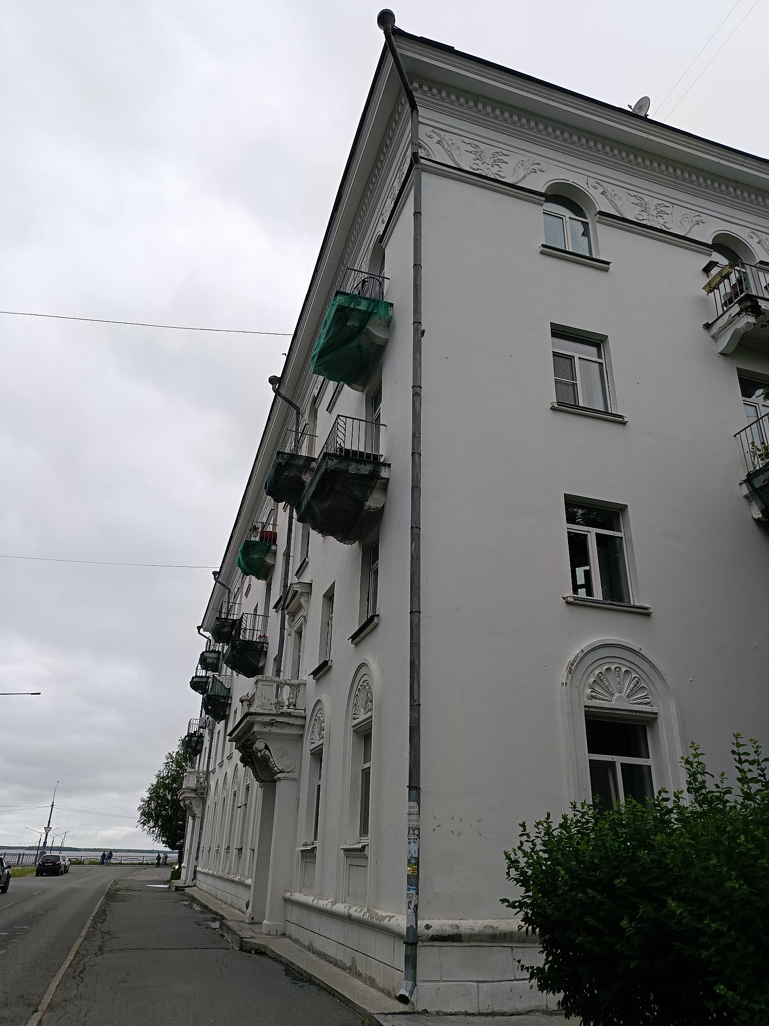 The image shows a white, multi-story apartment building with a distinctly European architectural style, featuring ornate detailing along the roofline and around the arched windows. Several balconies jut out from the building, some covered with green tarps, suggesting potential ongoing construction or maintenance. The building faces a street with a sidewalk and a body of water in the distance, likely a river or bay, given the location context of Arkhangelsk, Russia. The sky is overcast and gray, indicating a cloudy day, possibly with light rain or fog. A few cars are visible on the road, and a few people are seen walking along the waterfront promenade. The overall mood is somewhat somber due to the weather, but the grandeur of the building hints at a historical and established urban setting. No discernible text is visible in the image.