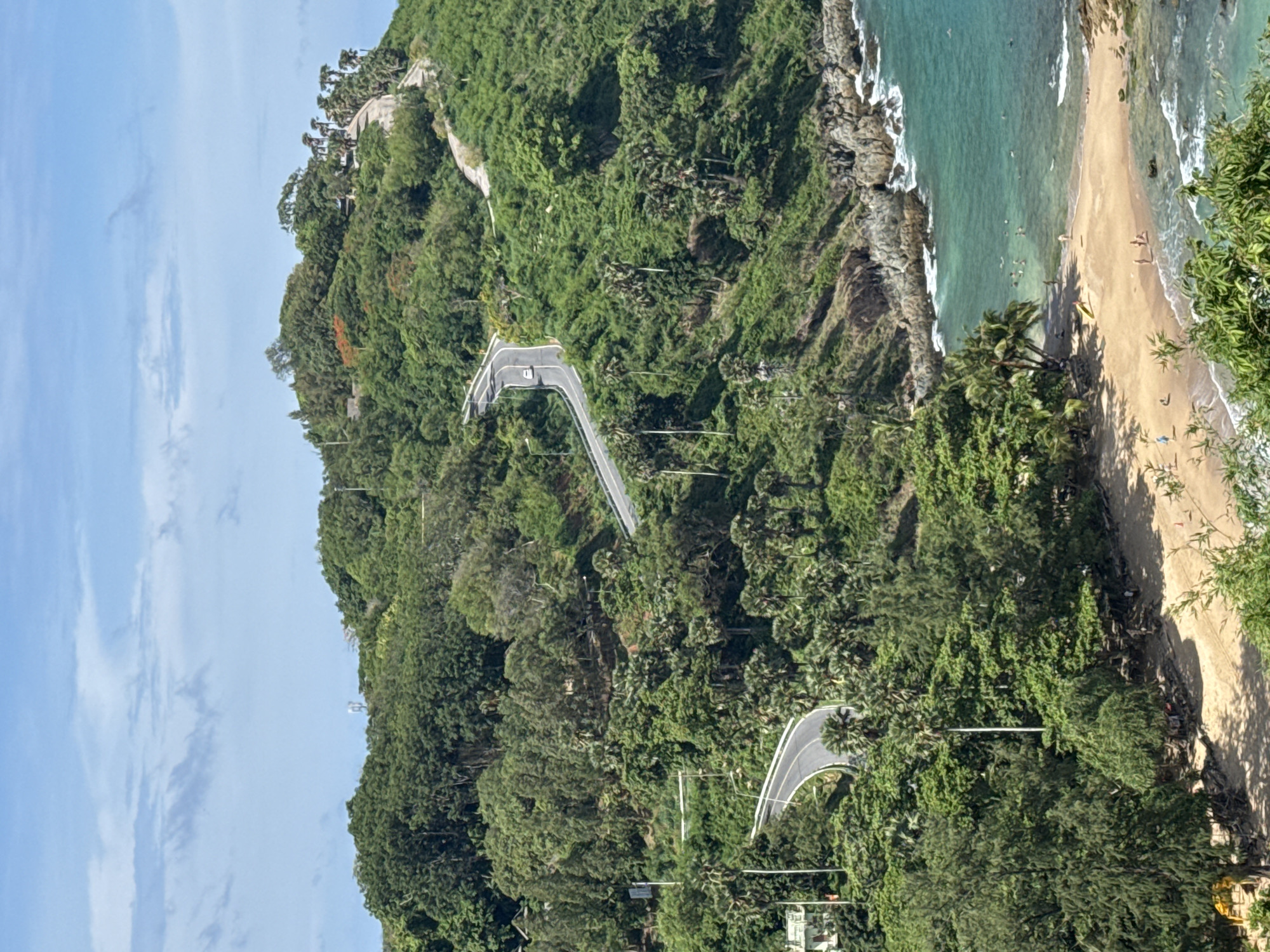 This is a high-angle, full shot of a scenic coastal road winding up a lush green, hilly landscape in Rawai, Thailand. The road, paved with asphalt, features white lane markings and curves dramatically through dense foliage dominated by various trees and palm trees. A single white car is visible on the road, ascending the hill.
Below the road, a small, sandy beach is nestled between rocky outcrops and the turquoise ocean. Several people are scattered on the beach and in the shallow water, engaged in activities such as swimming and sunbathing. A yellow kayak rests on the sand. The water is clear, revealing a rocky seabed in some areas, and waves gently lap the shore. The sky above is a clear blue with scattered white clouds. The overall impression is one of a tropical paradise with a well-maintained road providing access to the stunning natural beauty.