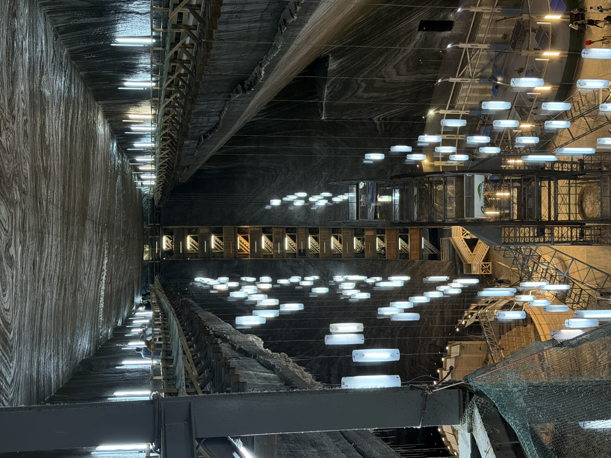 The image depicts the interior of the Turda Salt Mine in Boj-Cătun, Romania. The mine's rough-hewn walls are visible, illuminated by rows of fluorescent lights along the upper reaches. Suspended below are platforms and structures adorned with numerous horizontally-oriented light fixtures resembling glowing oblongs. Some platforms appear to have metal scaffolding. A green mesh netting is visible in the foreground. The mine's interior appears vast and multi-tiered.