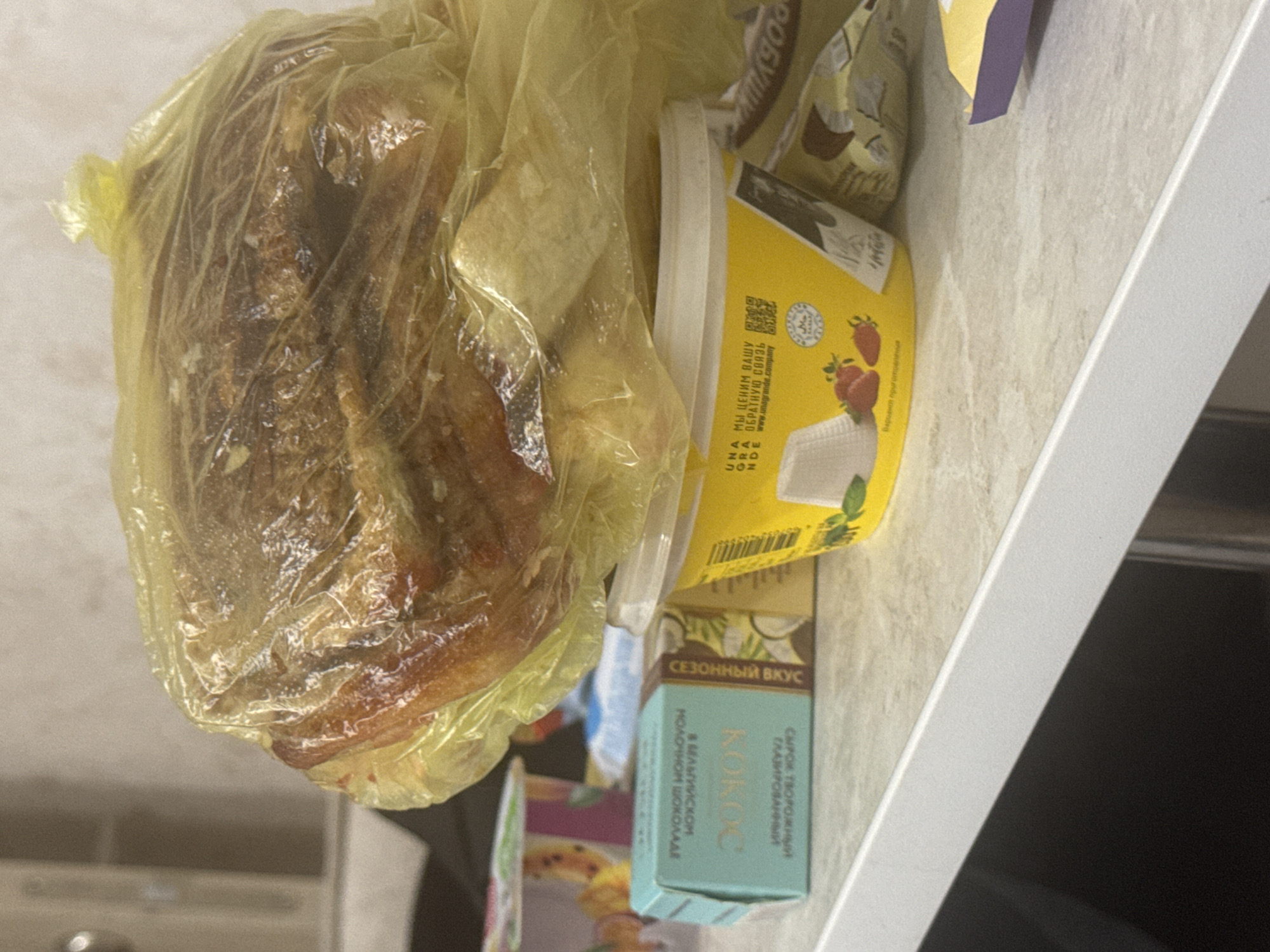 The image shows a close-up, eye-level shot of a white countertop with various food items. In the foreground, a portion of a dark surface is visible, possibly a table or appliance. The countertop itself has a speckled, light-colored texture.
Dominating the center of the frame is a large, baked item, possibly bread or a pastry, enclosed in a translucent yellow plastic bag. The item is golden brown and appears to be resting on a yellow plastic container.
To the left of the yellow container is a small, teal-colored box with Cyrillic text and an image of coconut. This box is positioned slightly behind and to the left of the yellow container. To the right of the yellow container, partially visible, is a brown and white patterned package, also with Cyrillic text.
The yellow container has text and graphics on its surface. Visible text includes Cyrillic script and the word "UNA". There are also images of strawberries. A circular emblem with "HALAL" written on it is present.
In the background, out of focus, are other food packaging items and a blurred lighter-colored surface, possibly a wall or cabinets. The lighting suggests an indoor environment, likely a kitchen.
There are no people visible in the image. No specific location in Saratov, Russia, can be determined from the visual evidence. The scene depicts food items on a countertop.