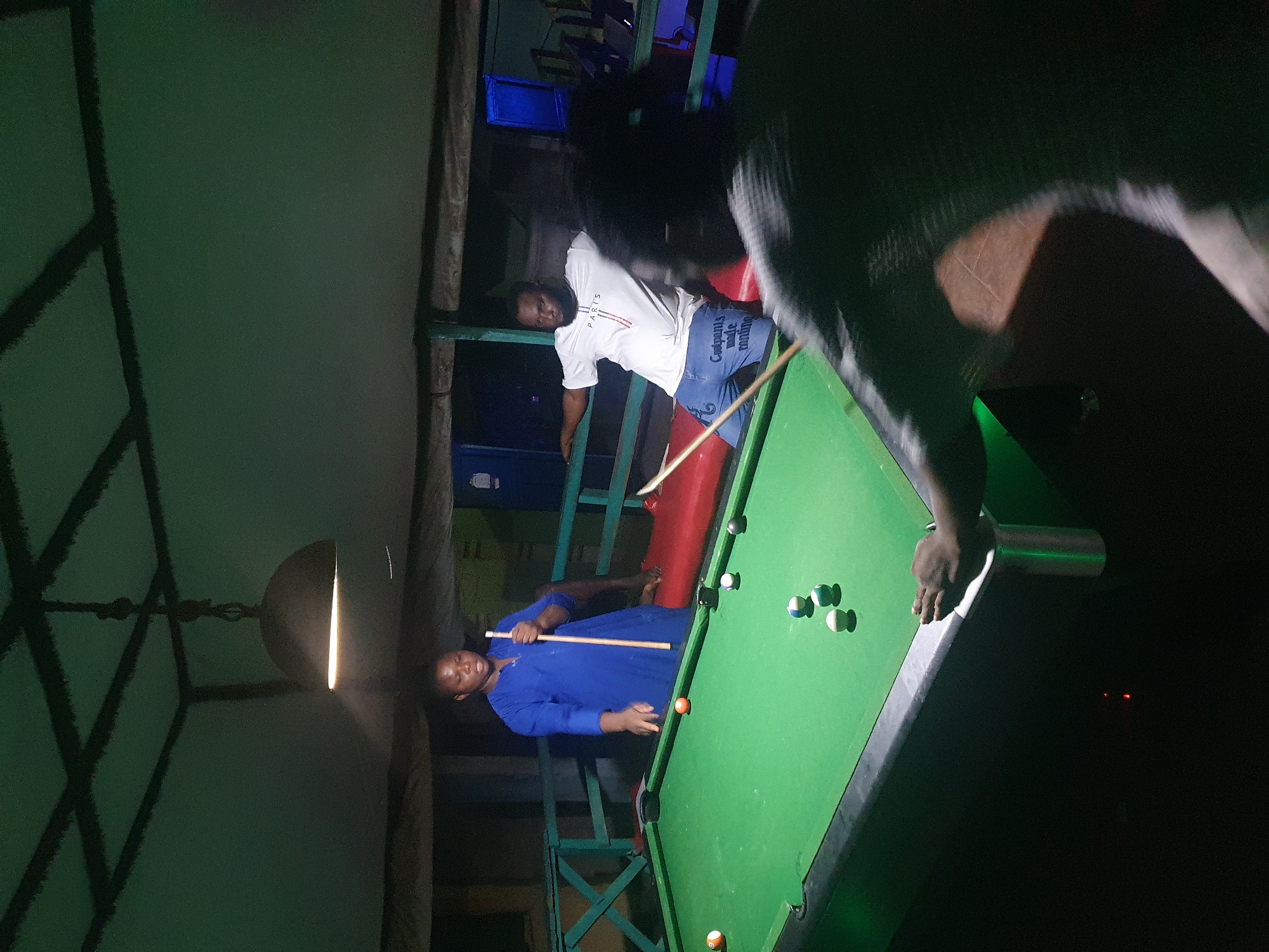 In Lokoja, Nigeria, a dimly lit indoor billiards hall, the scene captures three individuals engaged in a game of pool. A woman in a vibrant blue kaftan stands to the left, holding a cue stick and seemingly observing the table, with a slight smile on her face. To her right, a man in a white "PARIS" t-shirt and blue shorts leans on a railing, his posture suggesting a break from play. In the foreground, a third person, mostly obscured by shadow and out of focus, is partially visible, their hand resting on the edge of the pool table. The green felt of the pool table is scattered with several balls, including a prominent orange ball near the center. The environment is characterized by a low ceiling with exposed beams, and a hanging pendant light fixture that casts a warm glow. The walls appear to be made of wood and metal railings, suggesting a casual, perhaps outdoor-covered, setting.