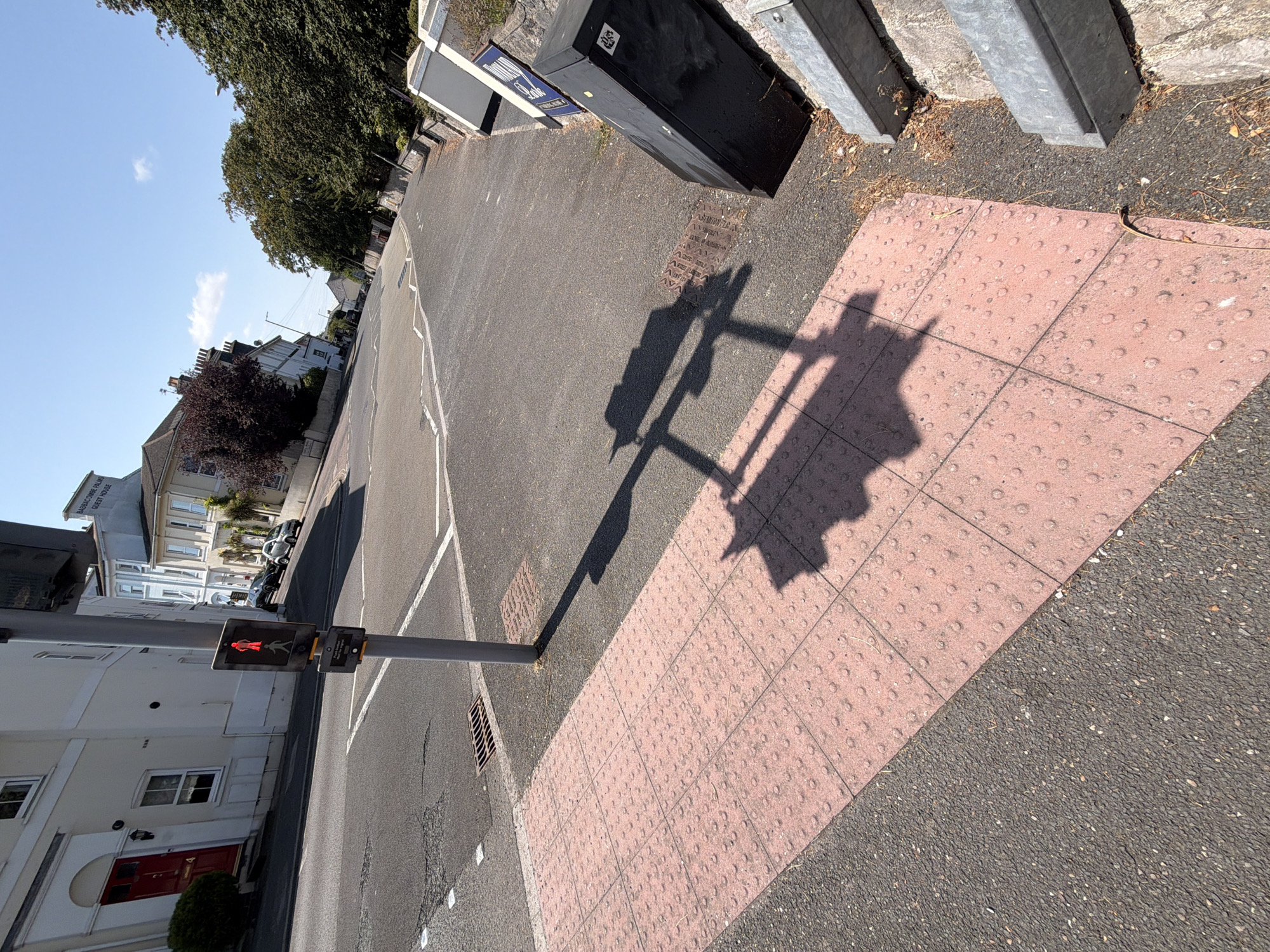 This image depicts a street scene in Torquay, United Kingdom, with a focus on a pedestrian crossing. A traffic light pole stands prominently, displaying a red pedestrian signal. The crossing itself is marked with tactile paving, characterized by rows of raised circular studs on pink tiles. The surrounding environment includes asphalt roads, a storm drain, and a sidewalk. In the background, residential buildings with white facades and red-tiled roofs are visible, along with mature trees and a clear blue sky. The sun casts long shadows, indicating a sunny day. The overall impression is that of a quiet urban street.