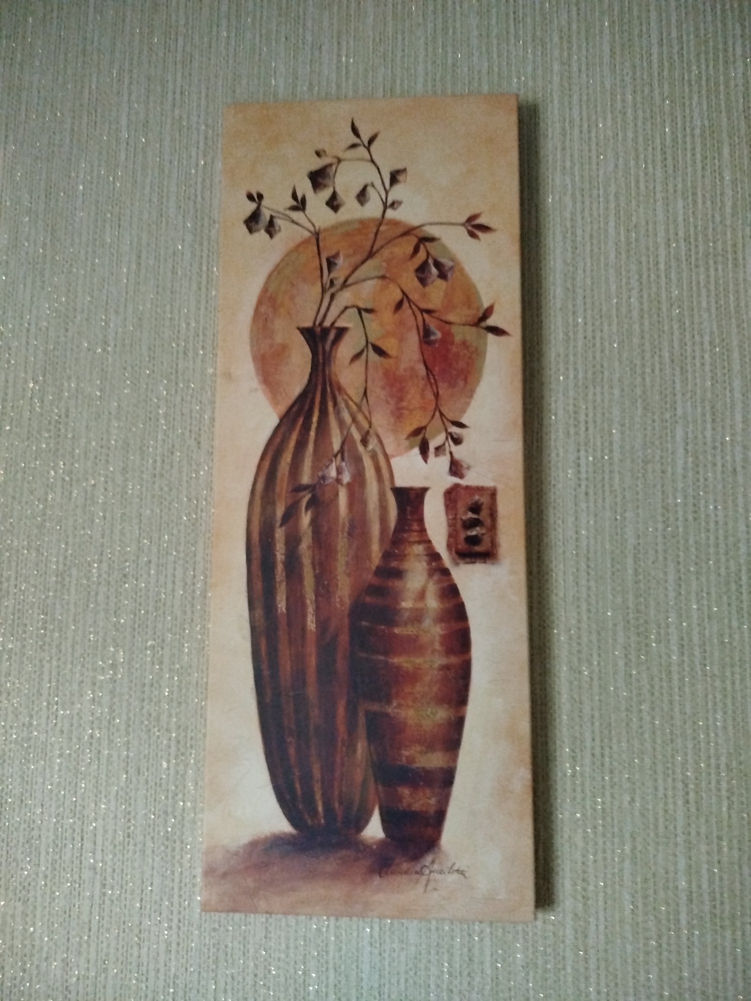 A vertical, rectangular painting is mounted on a textured, pale green wall. The painting depicts a still life of two ceramic vases. The larger vase, on the left, is dark brown with vertical ribbed detailing. It is filled with thin, brown branches that have dark, leaf-like shapes. Behind and to the right of the larger vase, a smaller, similarly decorated vase is visible. To the right of the smaller vase, there is a small, square, brown object with three dark circular elements. A pale orange, circular shape, possibly representing a sun or moon, is visible in the background of the painting, behind the upper portions of the vases and the branches. A faint, handwritten signature is present in the lower left corner of the painting. The wall has a subtle sparkle, as if embedded with fine glitter. The image is taken from a straight-on perspective, with the painting slightly off-center to the right.