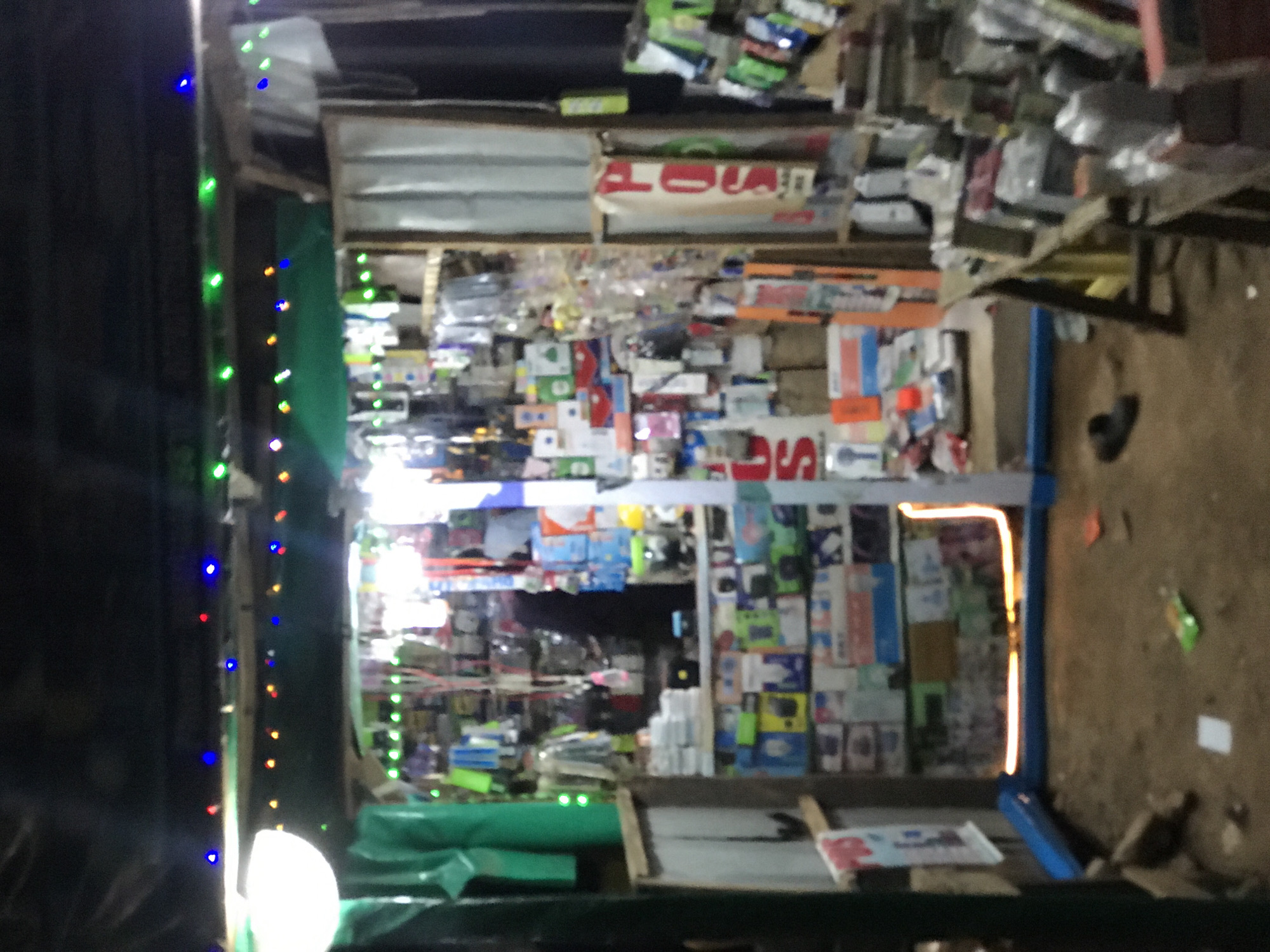 The image shows the front of a small shop in Galadimawa, Nigeria. The shop is packed with various items, seemingly electronics and accessories, densely arranged on shelves and walls. A string of colorful lights is strung along the shop's entrance. The ground outside is dirt. A blue metal frame partially outlines the right side of the shop's entrance. A single shoe lies on the ground near a table outside the shop.
