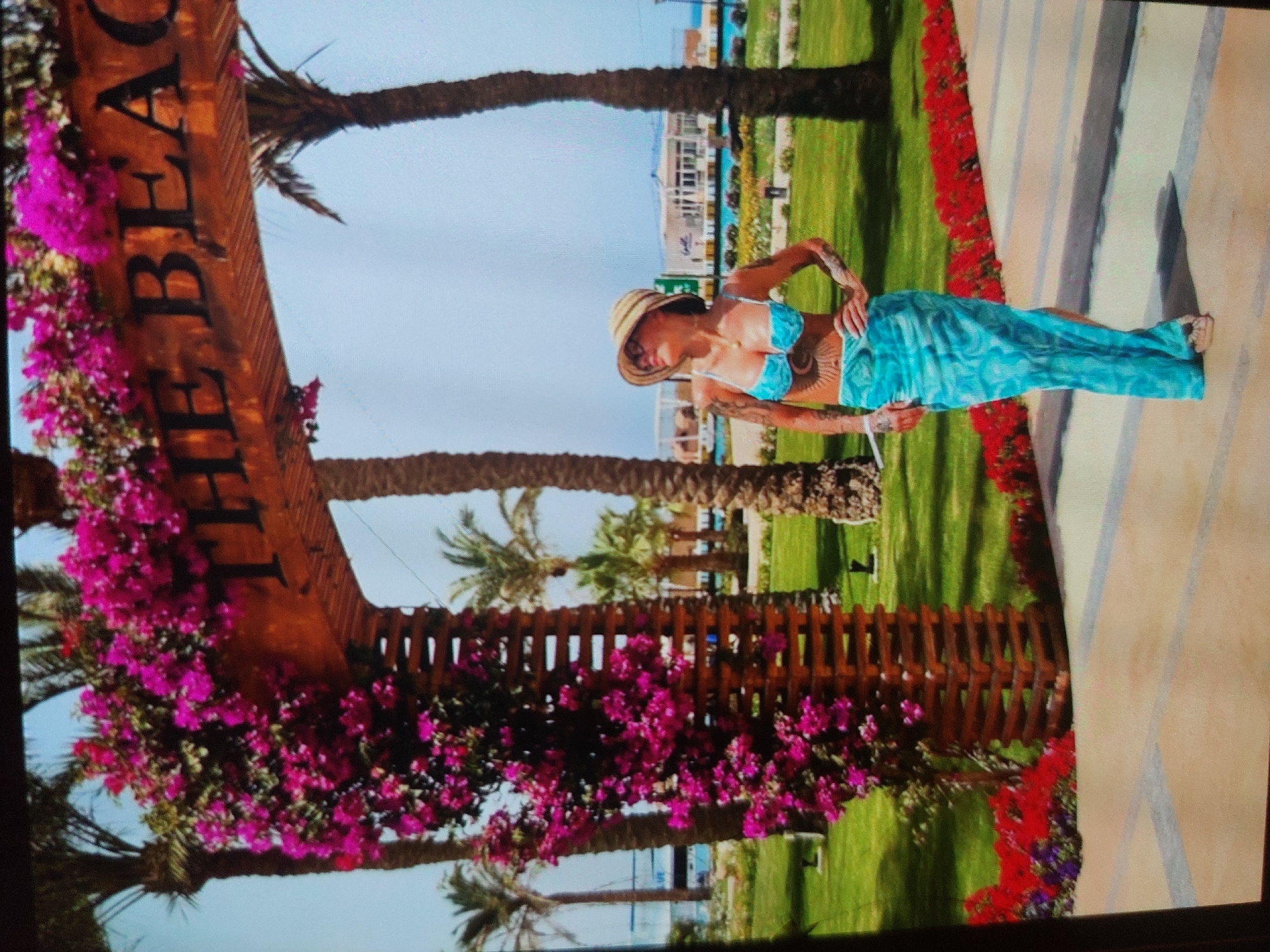 The image is rotated 90 degrees clockwise, showcasing a vibrant outdoor scene, likely at a resort in Tsarevo, Bulgaria, during a clear, sunny day.
The central subject is a woman posing for the camera, positioned slightly to the right of a prominent wooden archway. She is wearing a light blue bikini top with a swirling pattern and matching sheer, wide-legged pants or a sarong. Her attire is complemented by a straw hat and a gold necklace. Notable tattoos are visible on her arms and midriff. She stands with one hand on her hip, striking a confident pose.
On the left side of the frame, the large wooden archway is elaborately adorned with abundant, bright pink flowers, resembling bougainvillea. Dark letters spelling "THE BEACH" are clearly visible running vertically along the archway's wooden structure.
The background features several tall palm trees set against a clear, bright blue sky. Behind the trees, multi-story resort buildings with balconies are discernible. The foreground and midground consist of neatly manicured green lawns, bordered by rows of vibrant red flowers, and a light-colored paved walkway. The bright, even lighting throughout the scene indicates a mid-day setting with excellent weather, characteristic of a tropical or Mediterranean resort.