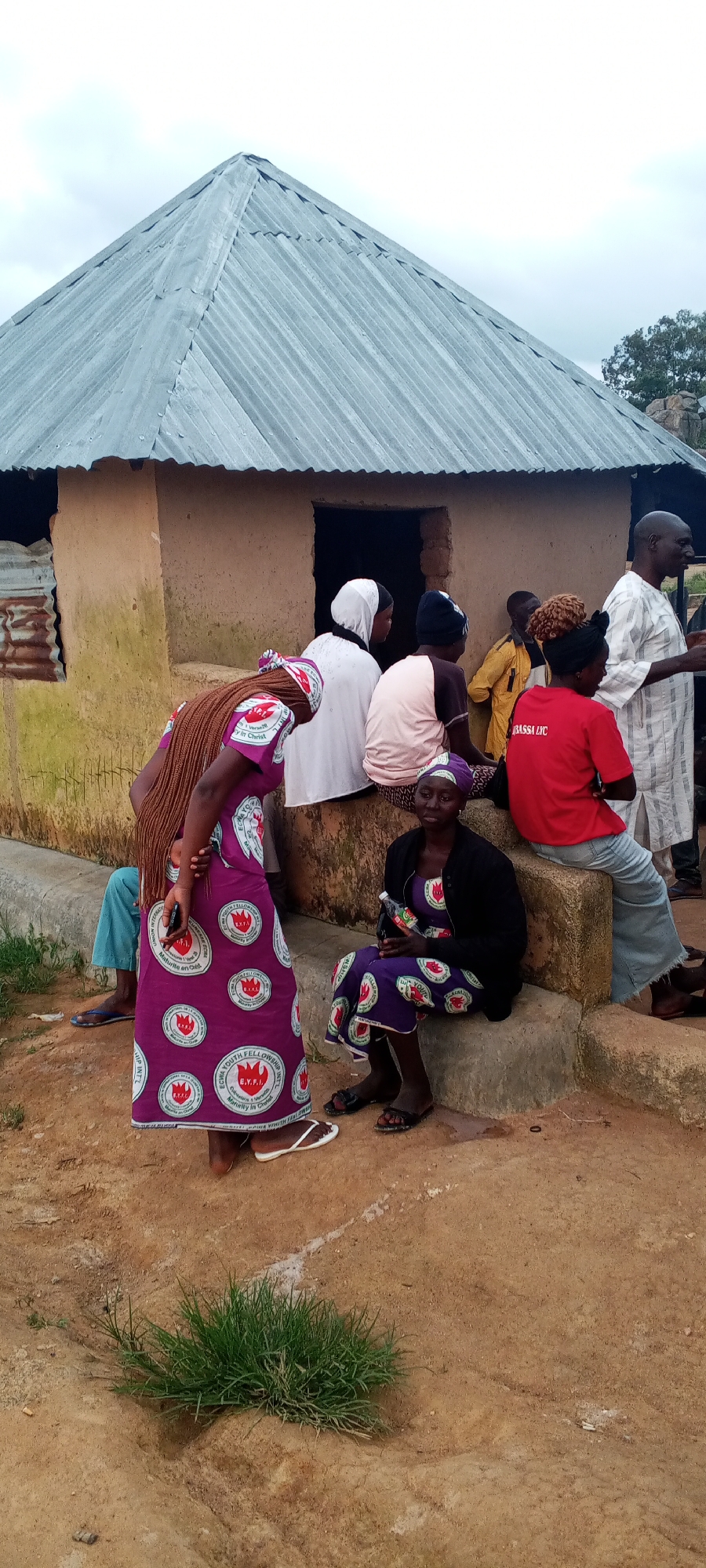 A group of people, some of whom are women wearing traditional African clothing, are gathered outside a rustic building with a corrugated metal roof. The building appears to be made of mud or adobe and has a dark, recessed doorway. One woman in the foreground, facing away from the camera, wears a long, purple dress with a repeating circular emblem and has long braided hair. Another woman, seated on the steps of the building, also wears a purple dress with the same emblem and a black jacket. A man in a white striped robe stands to the right, speaking to someone. The ground is dry and dusty, with patches of green grass. The scene appears to be in a rural or semi-rural setting. This image was taken in Ohit, Nigeria.