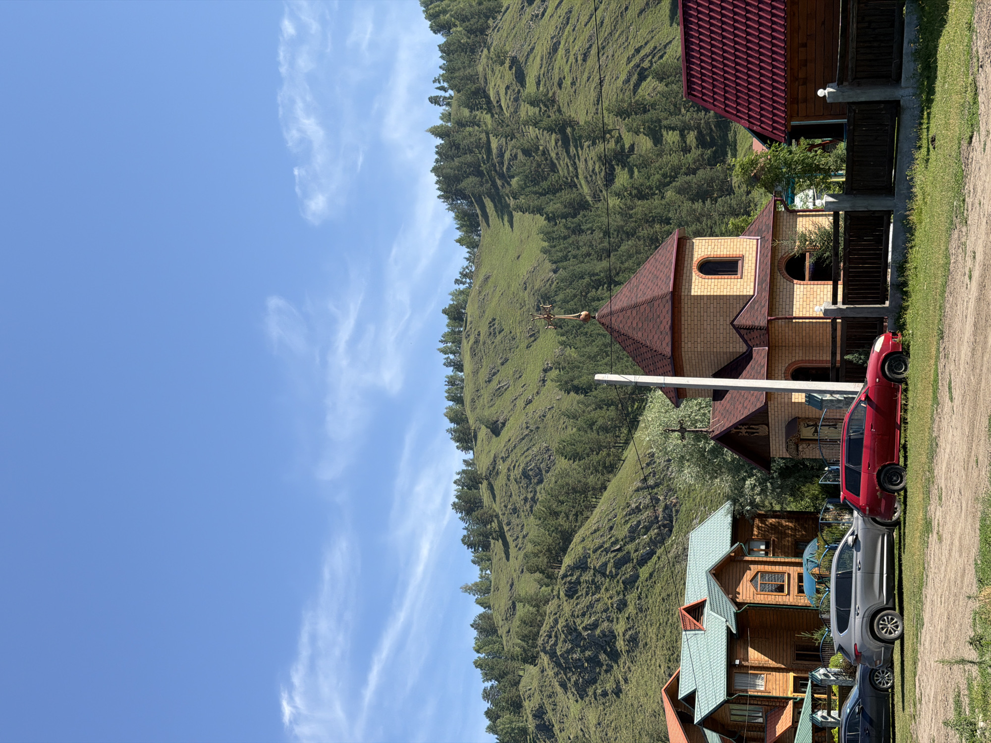 A bright, sunny day with a clear blue sky and sparse wispy clouds offers a view of a village nestled at the foot of a lush, green mountainside in Elekmonar, Russia. The mountainside is covered in a dense forest of evergreen trees, with some patches of grass and rocky outcrops visible. In the foreground, several wooden houses with distinctively colored roofs are clustered together. To the left, a multi-story wooden house with a teal roof is prominent. Next to it, a smaller wooden structure with a similar teal roof stands. In the center, a small, brick church with a reddish-brown tiled roof and a golden cross on top is visible, flanked by a wooden fence. To the right of the church, another wooden house with a dark red tiled roof is partially obscured by the church. In front of the houses and church, a dirt and grass area serves as a parking lot. Three cars are parked there: a dark gray sedan on the left, a silver SUV in the middle, and a red station wagon on the right. The scene appears peaceful and residential, with no people visible. The overall impression is one of a quiet, rural community set against a beautiful natural backdrop.