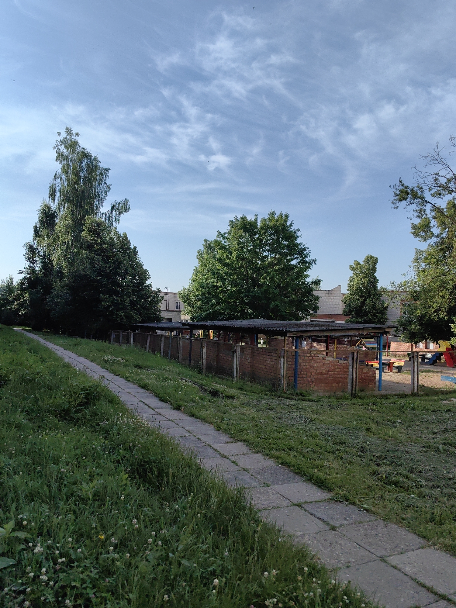 The image shows a view of a playground area on a sunny day with a clear blue sky and scattered white clouds. In the foreground, a concrete pathway runs diagonally from the bottom left corner towards the center of the image, bordered by tall green grass. Beyond the pathway, a low brick wall surrounds a structure with a corrugated roof, possibly a shelter or an unfinished building. In the background, there is a two-story building made of light-colored brick. To the right of the structure, a playground with colorful slides and benches is visible. Several trees with lush green foliage are scattered throughout the scene. The overall impression is of a recreational space within a residential or educational institution in Fanipal, Belarus. There are no people visible in the image.