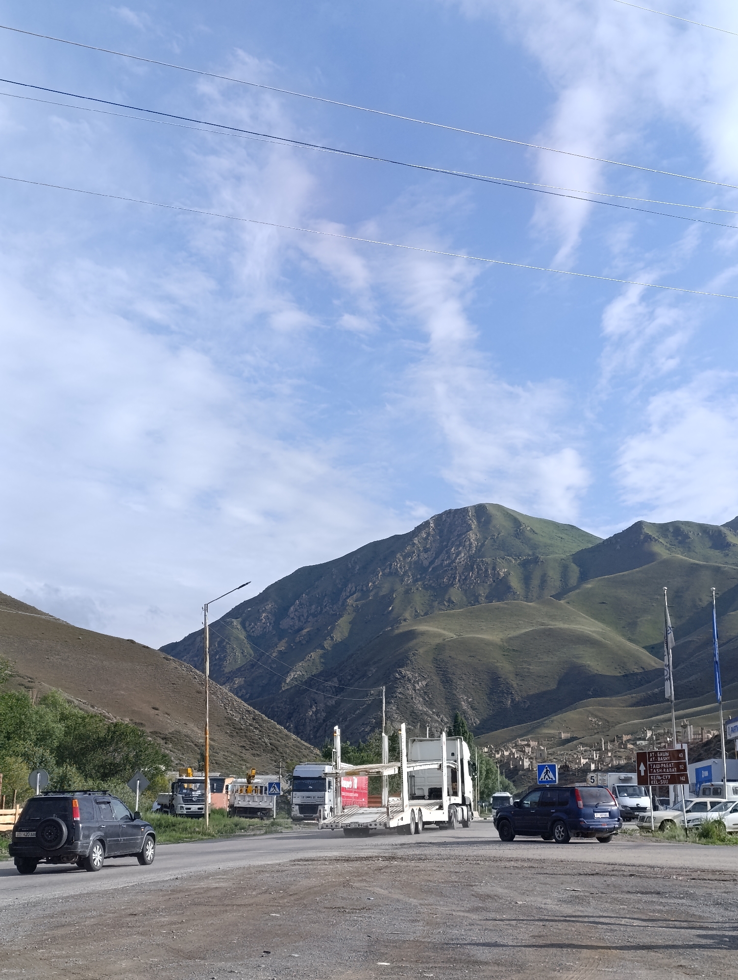 The image depicts a roadside scene in a mountainous region, likely near Naryn City, Kyrgyzstan. In the foreground, a gray asphalt road curves to the left. A black SUV is parked on the side of the road, facing away from the camera. Further down the road, several large trucks are visible, including a white car carrier truck. In the background, rolling green hills rise to meet a sky filled with scattered white clouds. A small village with stone buildings is nestled on the slopes of the hills. Several directional signs with Cyrillic lettering are posted near the road, indicating distances to various locations. A street lamp stands on a pole to the left of the frame. The overall impression is one of a rural, mountainous landscape with active road traffic.