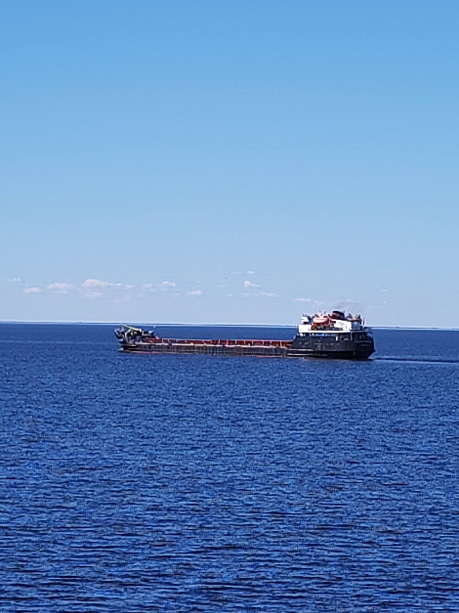 A long cargo ship, likely a self-propelled barge, is depicted traversing a vast expanse of calm, deep blue water under a bright, clear blue sky. The vessel is moving from left to right across the frame, leaving a subtle wake. Its hull is dark (black or dark grey), with a reddish-brown color visible in its open cargo holds, suggesting it is carrying bulk material.
The ship's superstructure is located towards the stern, featuring white and dark sections with windows and lifeboats/rafts visible on its upper deck. Although blurry, white text is visible on the hull of the superstructure; upon closer inspection, the word "МЕХАНИК" (MECHANIC) can be discerned, suggesting a Russian ship name, possibly part of "MEKHANIK KRIVOV".
The water surface exhibits gentle ripples, reflecting the clear sky. A distant horizon line separates the water from the upper two-thirds of the image, which is dominated by the unobstructed blue sky with only a few faint, wispy white clouds visible along the horizon. The bright illumination indicates a sunny daytime scene. The setting suggests a large lake or wide river, consistent with the location of Belozersk, Russia, on Lake Beloye.