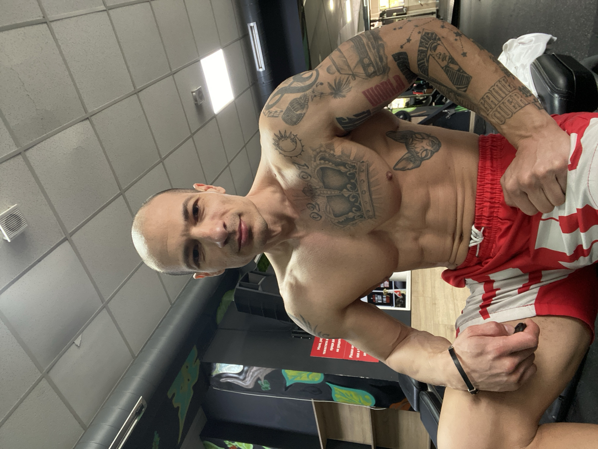 A muscular man with tattoos is posing indoors, likely in a gym, due to the presence of exercise equipment. He's wearing red and white shorts. The location is evidenced by a sign partially visible on the wall indicating it is located in the city of Pleven, Bulgaria.