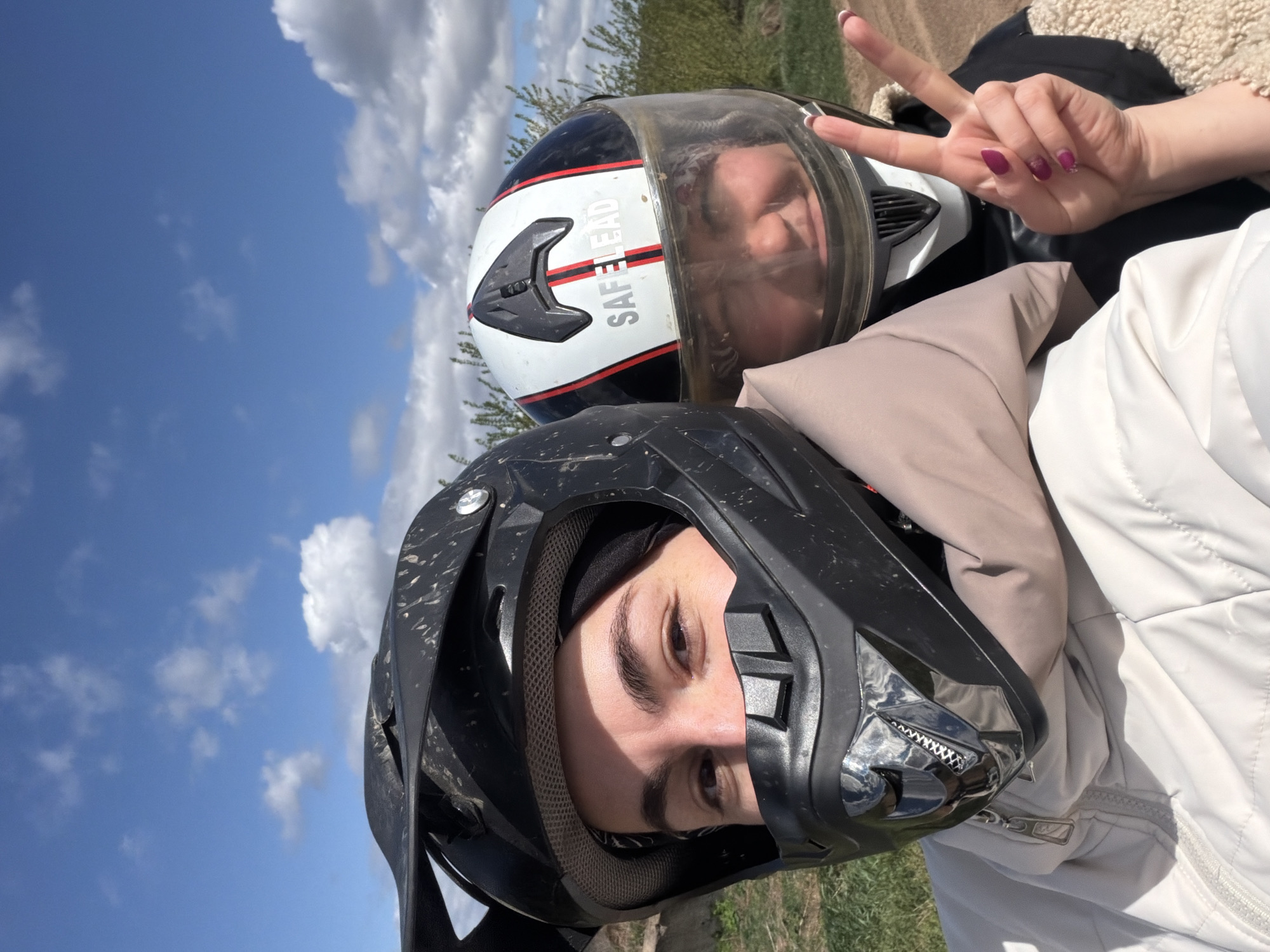 Two individuals are captured in an outdoor setting with a slightly elevated perspective, likely a self-taken photograph. The person closest to the camera wears a dark-colored, potentially dirt-covered helmet. The second individual is positioned behind, wearing a lighter-colored helmet with a clear visor. A hand displaying a peace sign is visible near the lighter-colored helmet. The backdrop consists of a blue sky with scattered clouds and some vegetation suggesting a rural or semi-rural environment. The image is rotated slightly, giving it a tilted appearance. The location in the city of Можга, Russia cannot be determined based on the visual information provided.