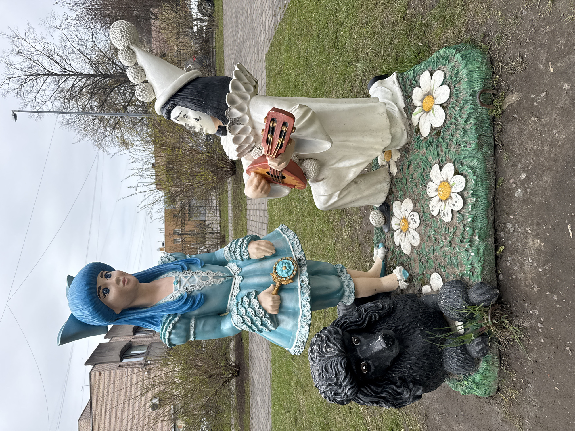 The image captures an outdoor scene featuring three colorful statues in what appears to be a park or public garden, set against an overcast sky.
To the left, a female figure stands, characterized by her vibrant blue, long wavy hair and a matching blue hat adorned with a bow. She wears a light blue dress with intricate white ruffled and laced details. In her right hand, she holds a small, ornate golden object with a blue centerpiece, possibly a mirror or locket. Her face is pale with striking large blue eyes, and she gazes slightly upwards.
Seated next to the blue figure is a black statue of a curly-haired dog, looking forward with dark, prominent eyes, its textured surface mimicking fur.
To the right, a male figure is depicted in a kneeling or sitting posture. He wears a tall, conical white hat topped with several white spherical pom-poms and a prominent white ruffled collar over a white costume, typical of a Pierrot character. His face is painted white with dark hair, and he holds a small, reddish-brown stringed instrument, resembling a lute or mandolin, positioned as if he is playing it.
Both humanoid figures are situated on distinct green bases. The female figure and the dog share a base featuring sculpted grass texture and painted white daisy-like flowers.
The setting includes a paved pathway, patches of grass, and several trees with bare branches, suggesting late winter or early spring. A multi-story building, made of brick or stone, is visible in the background, along with a streetlamp. The sky is uniformly gray and cloudy, indicating an overcast daytime. No text is visible in the image. The scene is located in Saint Petersburg, Russia.