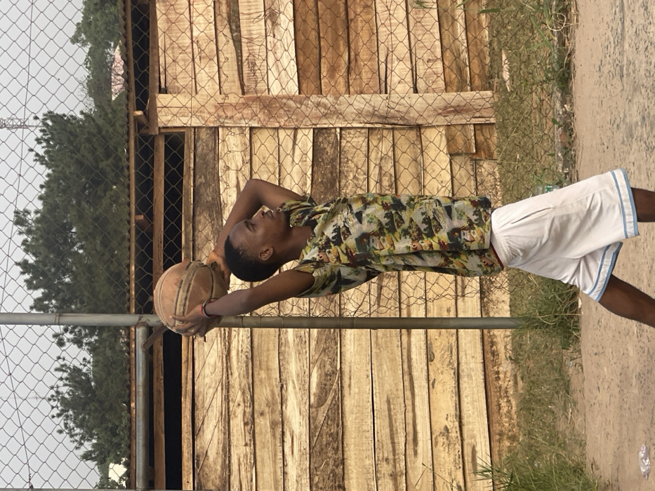 A young adult male stands in an outdoor setting in Umunekwu, Nigeria, positioned in profile facing right. He wears a multi-colored patterned short-sleeved shirt, white shorts with light blue trim, and a red wristband on his left wrist. Both hands hold a worn, brown basketball above his head in a shooting stance, arms bent at the elbows. Behind him is a chain-link fence, partially obscured by a metal pole, separating the foreground from a horizontal wooden plank structure in the mid-ground. Above the fence and wooden structure, green trees are visible against a hazy light gray sky. The ground consists of unpaved earth with scattered light green vegetation and a discarded clear plastic bottle in the lower-left.