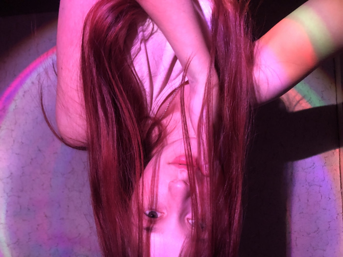 The scene is an indoor setting in the city of Фоки, Russia. A young female subject, with long, straight hair appearing reddish-purple due to strong artificial lighting, is positioned facing the camera. Her hair partially obscures the left side of her face. She has light skin and is wearing a light-colored garment, possibly a tank top or t-shirt, and a thin chain necklace. Her right hand is raised, with fingers resting near her chin and jawline. The background consists of a textured, light-colored wall, primarily dark except for two circular light projections. The projection on the left displays a rainbow spectrum, while the one on the right is predominantly pink and purple. The overall illumination of the subject and immediate environment is a vibrant pink and magenta.