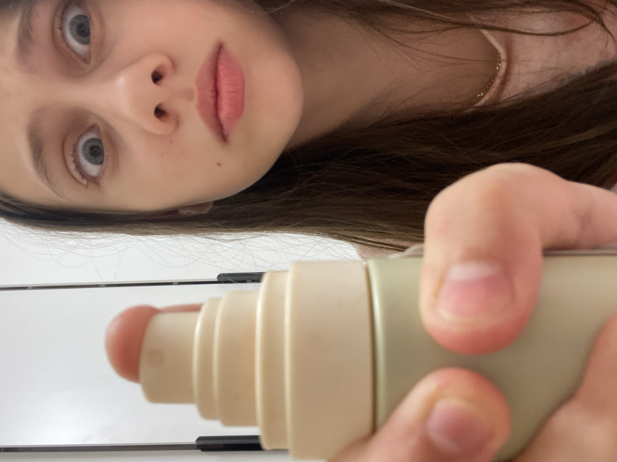 A young woman with light skin, grey eyes, and light brown hair is holding a cosmetic applicator in her right hand. The applicator is light beige with a rounded, pink-tinted tip. The woman's face is tilted towards the upper left corner of the frame. The background consists of white horizontal lines with dark accents, possibly a shelf or desk. There is no indication of the location being Izhevsk, Russia, or any other specific city.