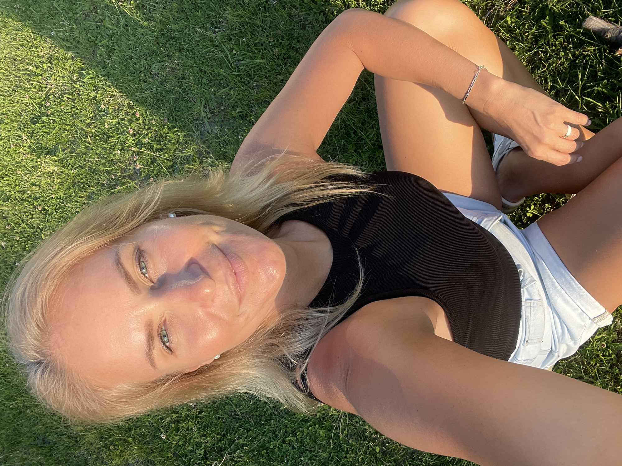 A blonde Caucasian female is depicted in a medium shot, crouched or kneeling on a patch of green grass under direct sunlight in what is identified as the city of Baranovka, Russia. She appears to be in her 30s or 40s, with blue eyes, and is smiling directly at the camera. Her blonde hair is parted slightly off-center. She is wearing a black ribbed tank top and light blue denim shorts. Adornments include small white pearl stud earrings, a silver link bracelet on her left wrist, and a white ring on her left ring finger. Her skin shows a tan complexion with highlights from the sun. The surrounding grass is green and contains small light-colored floral elements, possibly clover. Sunlight casts distinct shadows on the ground and illuminates the subject prominently.