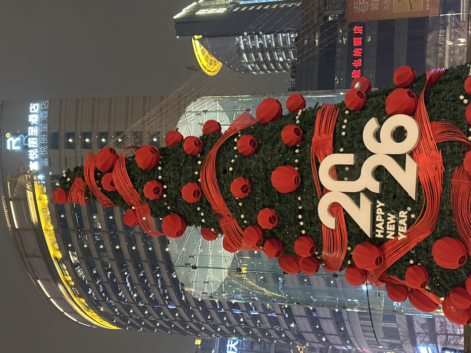 The image displays a large, elaborately decorated Christmas tree or festive display set against a backdrop of modern high-rise buildings at what appears to be night or dusk. The image is oriented vertically, rotated 90 degrees clockwise.
The central focus is a towering green tree, adorned with numerous red, spherical or lantern-like ornaments. These red decorations are arranged seemingly in clusters, and many are connected by slender red ribbons or strands that wrap around the tree. Small, white, bead-like lights or ornaments are also visible amidst the green foliage. A prominent feature on the tree is large, white numerical text "2026" followed by the words "HAPPY NEW YEAR" stacked vertically below the "26". The overall decoration style suggests a blend of traditional Christmas elements with Chinese New Year aesthetics, given the red lanterns.
In the background, several tall, contemporary buildings can be seen. To the left of the tree, a building with a lighted sign is visible. The sign, when rotated for readability, shows Chinese characters which the OCR identifies as "富悦丽呈酒店" (Fuyue Licheng Hotel). Another part of the sign appears to be "R". Further up and to the right, another building with glowing windows and what looks like a golden, round architectural feature or light is present. To the right of the tree, partially obscured, is another building with a red lighted sign in Chinese characters, identified by OCR as "维也纳酒店" (Vienna Hotel) with additional characters "春" and "养出". The buildings feature reflective surfaces and numerous windows, with some internal lights visible, contributing to the urban night scene.
The lighting in the image suggests either late evening or night, with artificial lights from the tree decorations and the buildings providing illumination against a dark, overcast sky. The atmosphere is festive and urban.