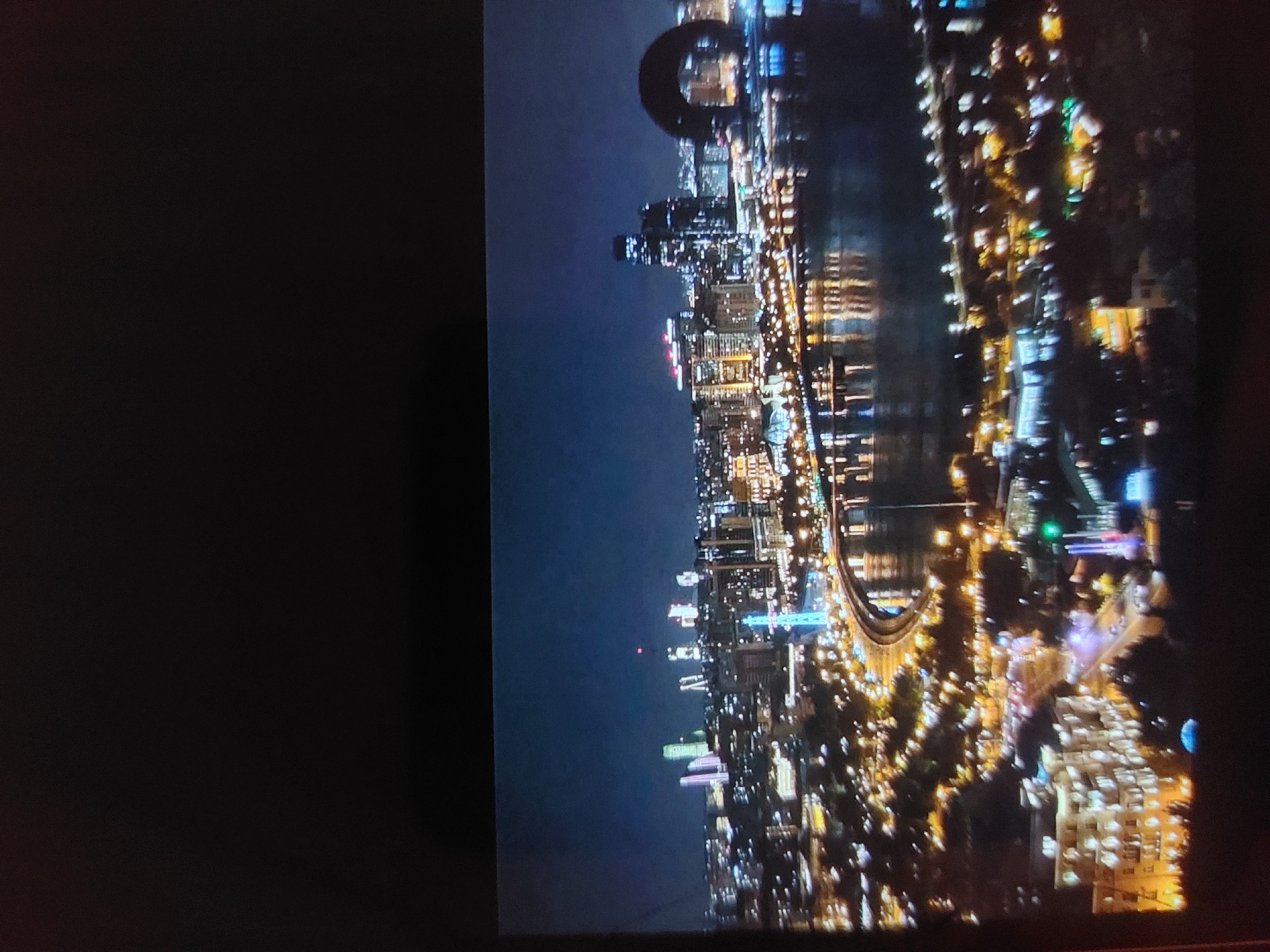 The image displays a vibrant night-time cityscape, oriented vertically with the city to the right. The scene features a multitude of brightly lit buildings, forming a modern skyline along a body of water, likely a river or bay, which occupies the lower and central parts of the frame. The water beautifully reflects the city lights, creating blurry, elongated streaks of illumination. A prominent bridge or elevated roadway, also aglow with lights, traverses the scene.
Among the numerous buildings, several skyscrapers are visible. A particularly distinctive feature is a large, dark, ring-like structure or arch silhouetted against the dark blue night sky, positioned high above the city, suggesting a significant architectural landmark. The sky is dark and clear, indicating a calm night. No people or discernible text are visible. The overall impression is a bustling metropolis captured during its evening hours, radiating urban energy. While the provided context suggests Tsarevo, Bulgaria, the extensive modern architecture and unique circular structure are more indicative of a larger, contemporary metropolitan area.