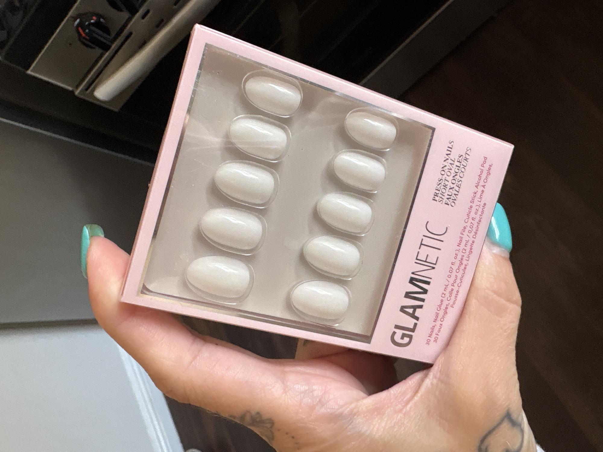 A hand holds a pink box of "Glamnetic" brand press-on nails with a clear plastic front showing ten oval-shaped, white nails. The package lists details in English and French, including the quantity of nails and the inclusion of nail glue, a file, cuticle stick, and alcohol pad. The hand holding the nails has a teal-colored fingernail and a tattoo is visible on the hand. The background includes an oven and dark-colored cabinetry. The scene appears to be indoors, possibly in a kitchen. The location is Seal Beach, United States of America.