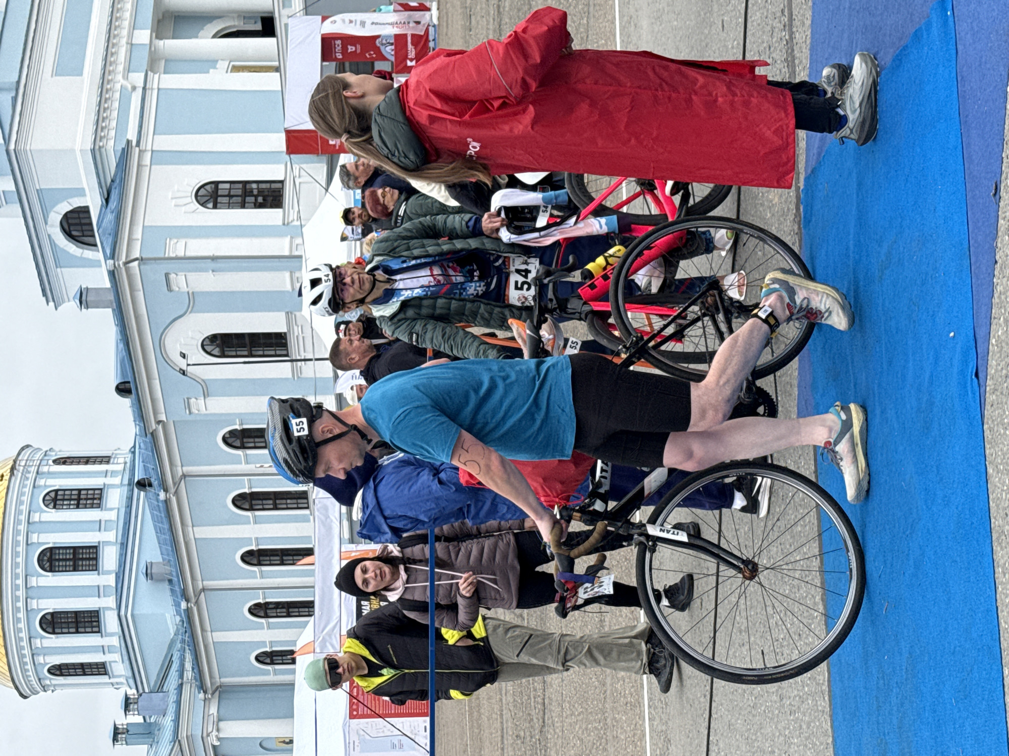 A group of people are gathered outdoors in front of a pale blue and white building with a golden dome, identified as the Votkinsk Cathedral of the Epiphany in Votkinsk, Russia. The foreground is dominated by a blue carpet laid out on asphalt. Several individuals are present, some with bicycles.
In the immediate foreground, a man in a turquoise t-shirt and black shorts is walking with a bicycle. He is wearing a helmet with the number 55 visible. To his left, another man wearing a green beanie and sunglasses stands next to a bicycle. Behind him, a woman in a dark puffy jacket and a black beanie is looking towards the man with the bicycle. Further left, a person in a blue jacket is partially visible.
To the right of the man in the turquoise shirt, a person wearing a green jacket with the number 54 visible on their chest is standing next to a pink bicycle. This person is also wearing a helmet. To the right of the pink bicycle, a person wearing a bright red hooded jacket is facing away from the camera.
In the background, more people are visible, as are tents and banners, suggesting an event is taking place. The building in the background exhibits classical architectural elements with arched windows and columns. The sky is overcast.