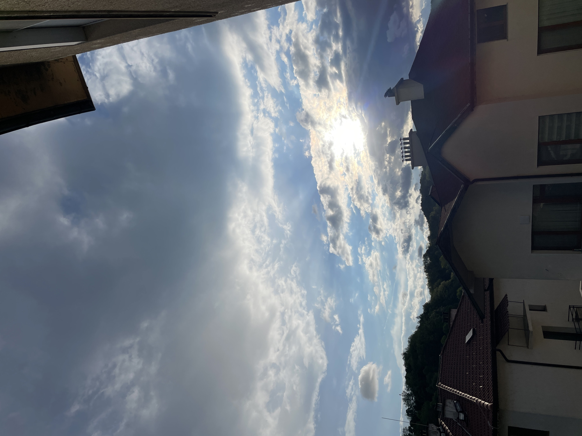 The image captures a daytime scene in Zlatograd, Bulgaria, featuring a sky filled with a mix of dark gray and white clouds, with the sun shining through a break in the cloud cover. The view is angled upwards, showing rooftops and the upper portions of several buildings.
The building on the right has a sloping red tile roof, white exterior walls, and visible windows and balconies. Adjacent to this building, a treeline suggests a nearby wooded area. On the left, a section of another building is visible, showing a rough, gray exterior. The sky dominates the majority of the frame.