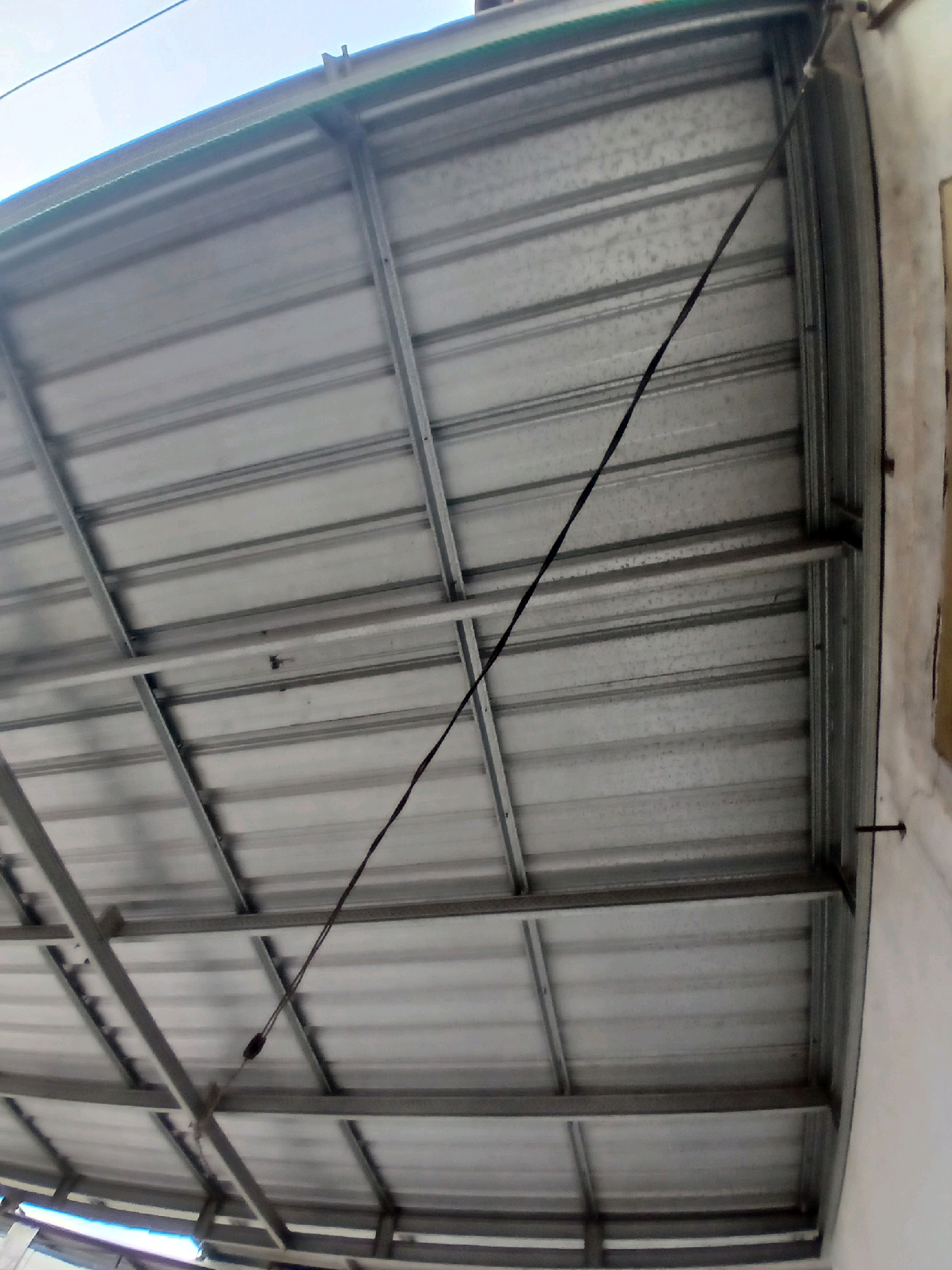 The image shows the underside of a corrugated metal roof supported by a metal frame. The roof is grey and has a ribbed texture. The supporting frame consists of several parallel metal beams and crossbeams. A thin black wire runs diagonally across the roof structure, secured at one point by a knot. The right side of the image shows a portion of a white wall, with a screw or nail protruding from it. The scene appears to be outdoors, with a bright sky visible at the top left. The location is identified as Tenggarang, Indonesia.