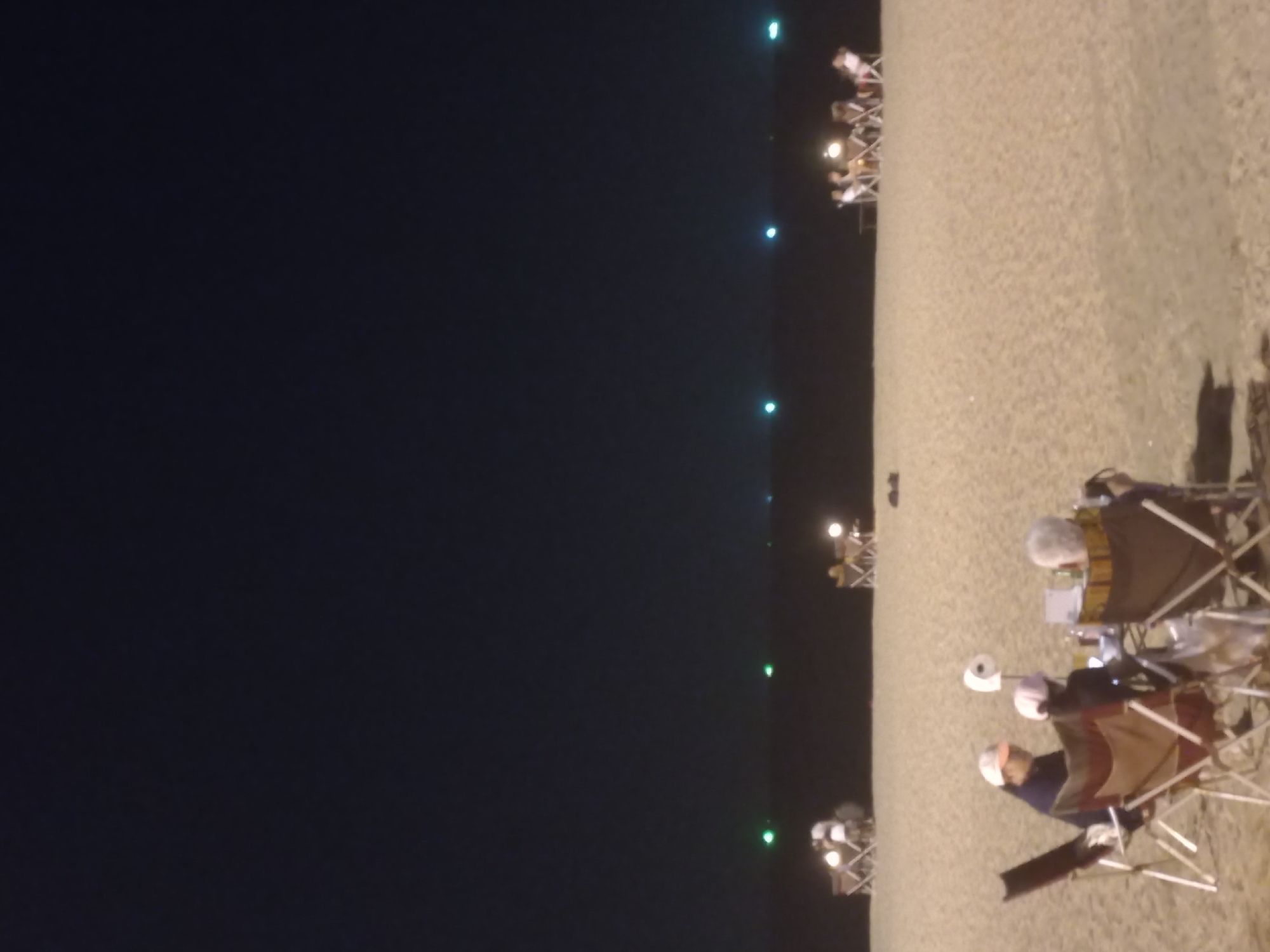 The image depicts a nighttime scene on a sandy beach, consistent with a coastal location like Pattaya City, Thailand. The photograph is oriented vertically, with the dark expanse of the ocean or sky occupying the left side, and the lighter-colored sand of the beach on the right.
Several groups of people are scattered along the beach, enjoying the evening. In the foreground, two individuals are clearly visible sitting on portable folding chairs. One person wears a light-colored hat or head covering, and various personal items are placed around them. Further down the beach, more groups are seated in similar chairs, some gathered around small, portable tables illuminated by their own light sources, such as lanterns or small lamps. These individual lights create pockets of warmth and intimacy on the otherwise dark beach.
In the distance, across the dark water or sky, several faint green and blue lights are visible, possibly indicating distant boats, buoys, or lights from the far shore. The activity appears to be one of relaxation and socializing, with people enjoying the nighttime atmosphere by the sea. The time of day is clearly night, with artificial lights being the primary illumination. No specific weather conditions are discernible, but the outdoor gathering suggests calm conditions. There is no visible text in the image.