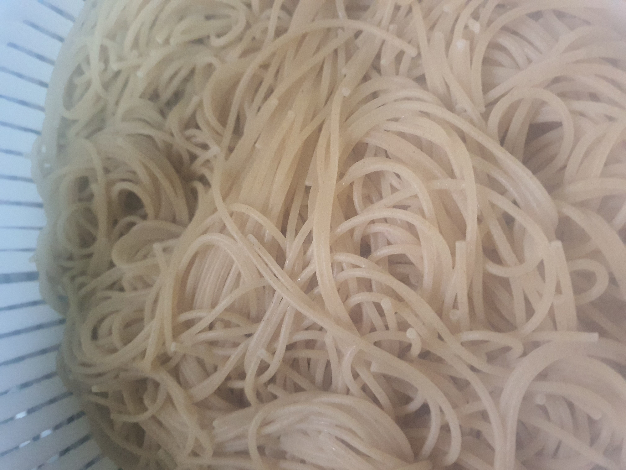 A close-up, overhead shot shows a pile of cooked spaghetti noodles in a white colander. The noodles are light brown and appear to be whole wheat. They are tangled and piled high, with some strands coiled into loops. The colander's white plastic bars are visible in the background at the top of the image. The lighting is diffused and even, suggesting an indoor setting. There are no people or other objects visible in the scene.