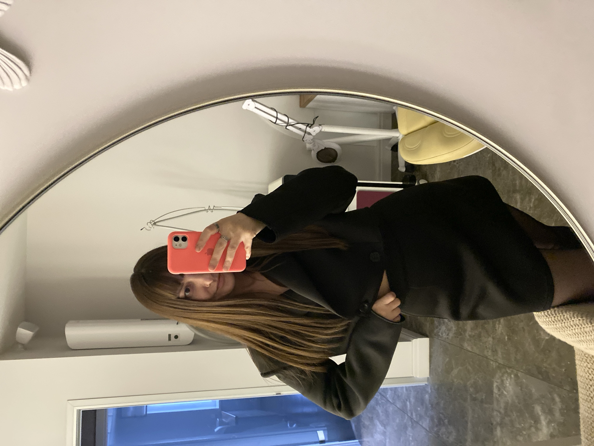 A mirror selfie, rotated 90 degrees clockwise, shows a person, likely female, with long, straight brown hair and bangs. The subject is wearing a black blazer or jacket and dark hosiery. She holds a coral-colored smartphone, possibly an iPhone given its dual camera lenses, in her right hand, with multiple rings visible on her fingers. Her face, reflected in the mirror, shows a neutral to slight smile, looking directly at the camera.
The central element is a large, round or oval mirror with a thin, light-colored frame, reflecting the person and the room. In the background reflections, parts of an indoor setting are visible: a pale yellow chair with a rounded, padded back, a white multi-jointed lamp or piece of equipment, a white rectangular device (possibly an air freshener or diffuser) placed on a shelf, and a white air conditioning unit or vent on the wall. The floor appears to be dark, possibly tiled or marbled. A light-colored, textured woven fabric, perhaps a blanket or rug, is also visible. The lighting suggests an indoor setting, and no specific time of day is discernible. No legible text is present in the image. This scene was captured in Votkinsk, Russia.