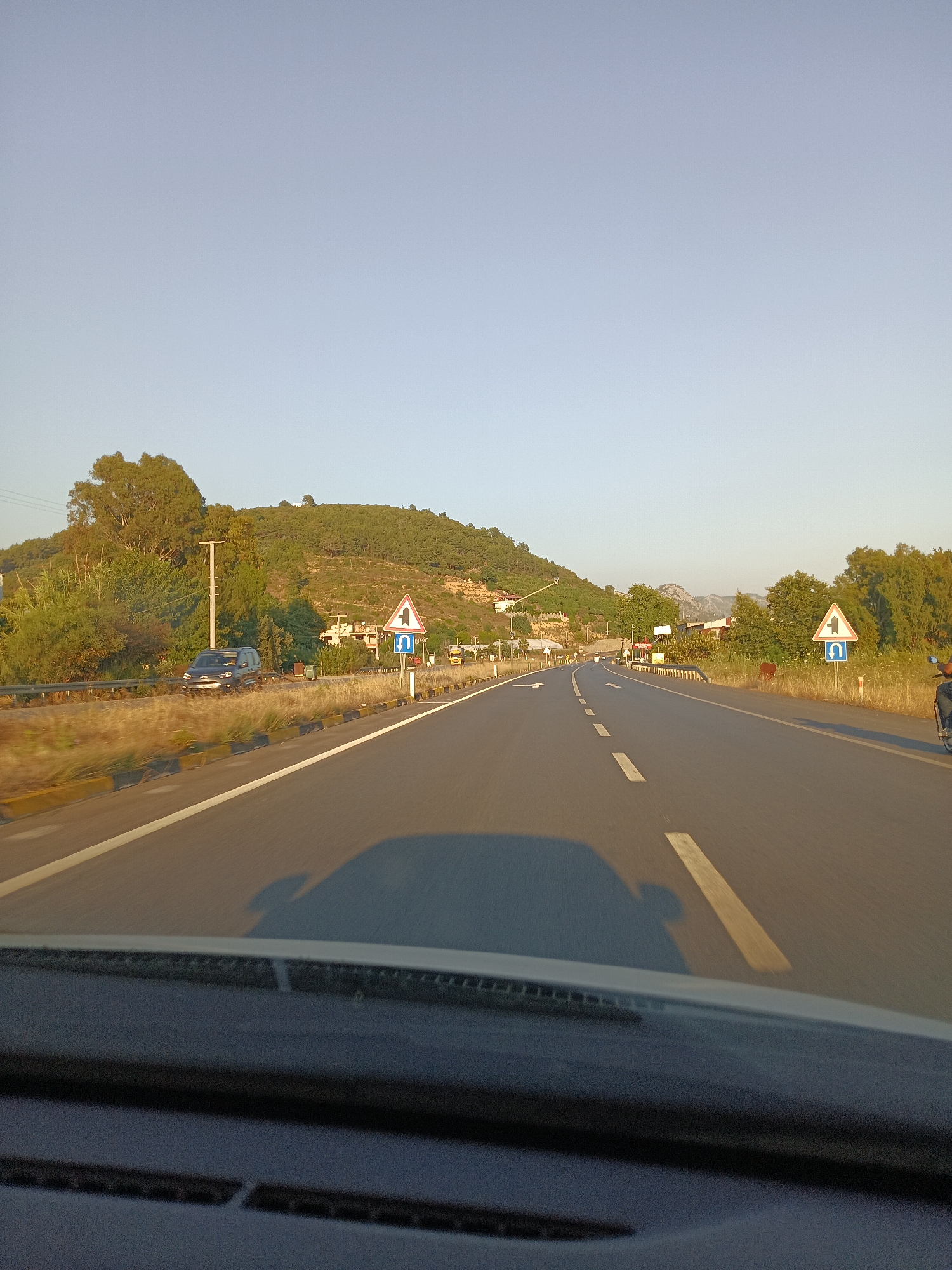 The image shows a view from a car driving on a highway. The road has multiple lanes and is bordered by dry grass and trees. On the right side of the road, there is a traffic sign indicating a sharp turn to the left and a U-turn. In the distance, there are hills covered with trees and some buildings. The sky is clear and blue, suggesting it is daytime. The scene appears to be in a rural or suburban area, likely outside the main city center of Alanya, Turkey, given the presence of a highway and less dense development. There is a dark-colored SUV in the left lane and a motorcycle rider partially visible on the right side of the road.
