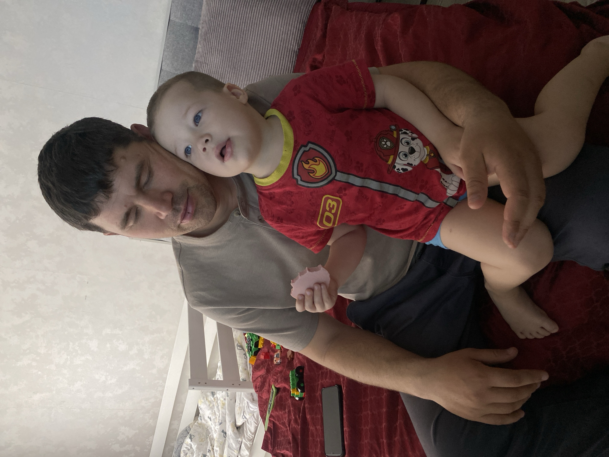 This image captures an indoor scene in Sochi, Russia, depicting an adult male and a young child seated together. The adult male, with short dark hair, wears a light tan short-sleeved t-shirt and dark blue trousers, positioned on a dark red textured blanket. He looks downwards with partially closed eyes, his right arm around the child. A small lesion or mark is visible on his upper lip. The child, a toddler with light brown hair and blue eyes, is seated on the man's lap, leaning against him. The child wears a red short-sleeved t-shirt featuring a "PAW Patrol" fire chief design (including a flame badge, the number '03', and Marshall the dog), with blue underwear visible. The child looks upwards and slightly to the right with a slightly open mouth, holding a small, light pink, amorphous object in their left hand.
To the left of the man's leg, a black smartphone rests on the red blanket. Behind the individuals, various colorful plastic toys, including green and yellow building blocks, are scattered on the red blanket. A white wooden bed frame, likely part of a bunk bed, is partially visible in the left background, featuring a white and yellow patterned duvet. A striped gray cushion is positioned to the right. The background wall is light-colored with a subtle textured pattern.