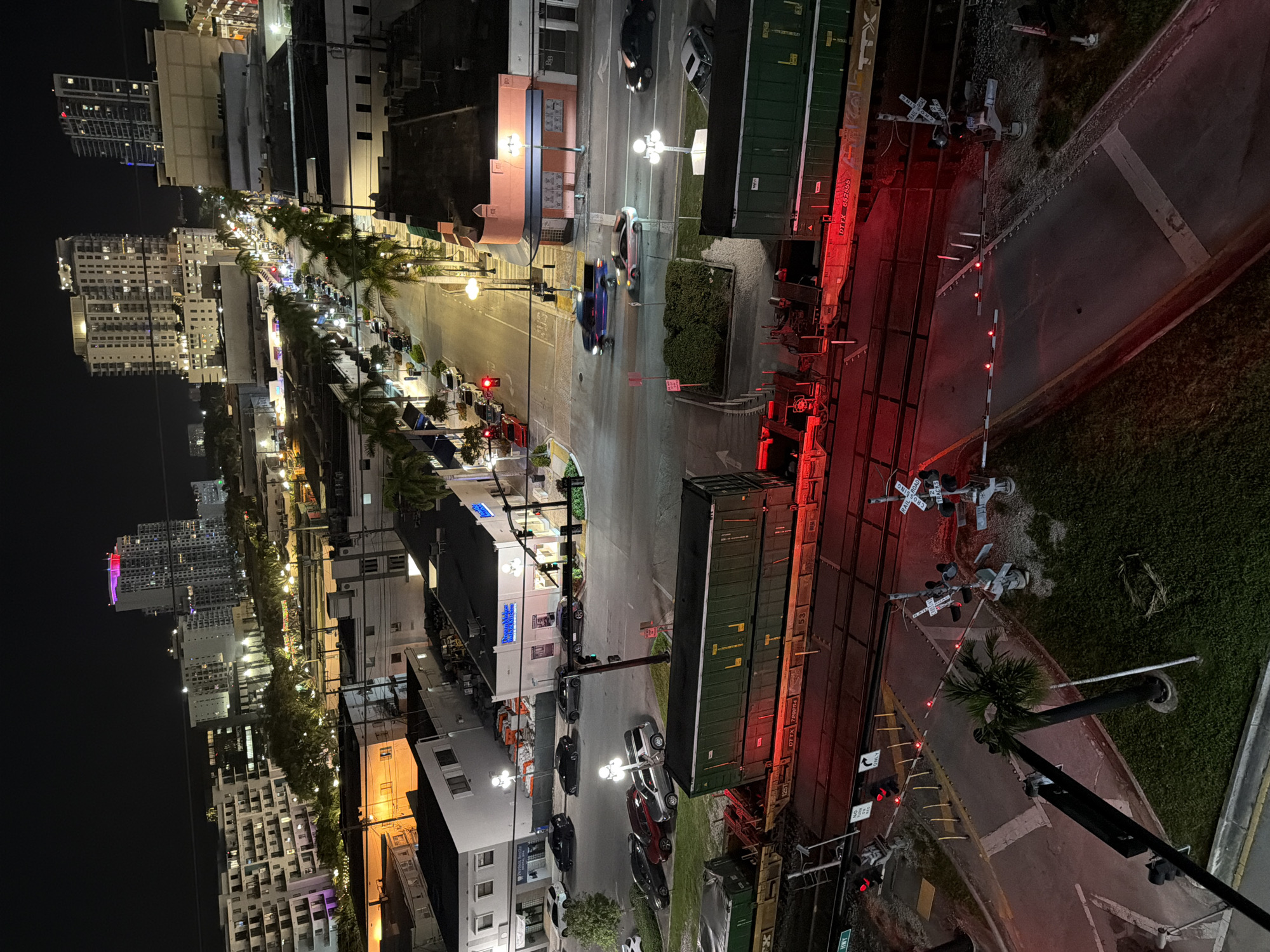 This is an aerial, nighttime photograph taken in Hollywood, Florida, USA. The scene captures a street with car traffic and a railroad crossing in the foreground. Several multi-story buildings, some illuminated and appearing to be apartments or hotels, are visible in the background, along with city lights and palm trees lining the street.
In the foreground, a train with several green and brown freight cars is stopped at a railroad crossing. The crossing gates are down, and red lights are flashing, indicating an approaching or present train. Cars are stopped on the street on either side of the tracks, waiting for the train to pass. The street itself is illuminated by streetlights and the lights from nearby buildings.
The buildings in the background vary in height and architectural style. Some have illuminated windows, suggesting occupancy. The overall impression is of a lively urban environment at night. The photograph is taken from a high vantage point, possibly from a taller building overlooking the street and railroad.