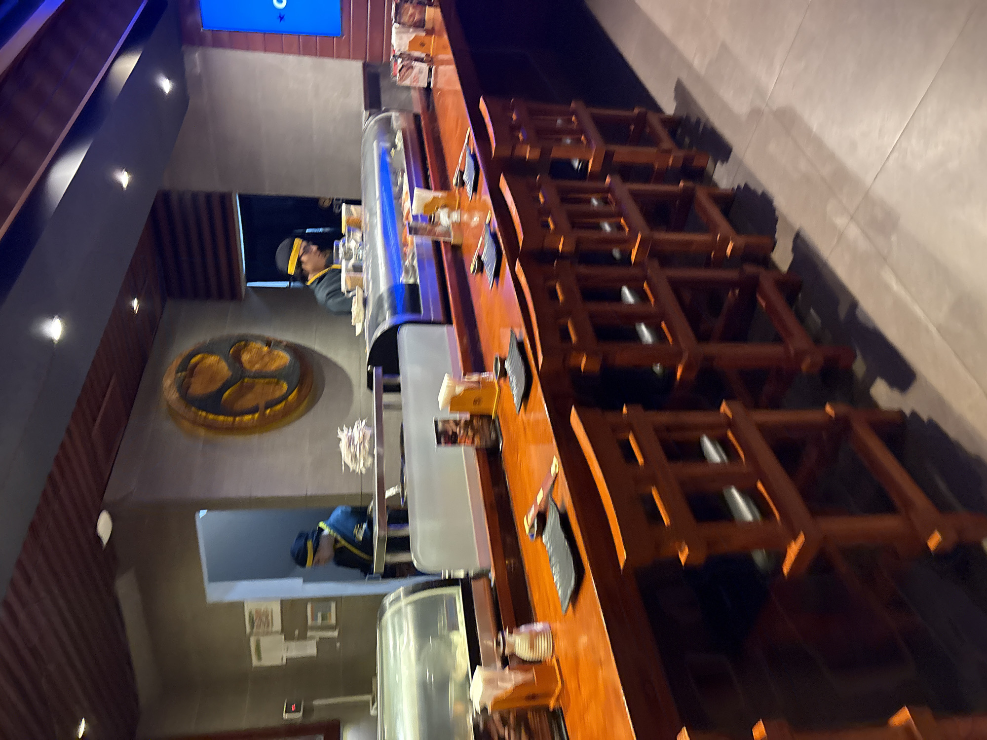 The image depicts the interior of a sushi restaurant in Magdalena del Mar, Peru. A wooden bar with a polished, orange-toned surface runs horizontally across the frame. Several wooden bar stools with an open, ladder-back design are positioned in front of the bar. On the bar, various items are visible including black rectangular plates, chopsticks, small white containers, a salt shaker, and paper napkins. Behind the bar, a refrigerated display case filled with sushi is prominent. Two individuals, presumably chefs or staff, are seen working behind the bar. One is wearing a blue uniform and a yellow headband, and is partially visible through a doorway. The other, also in a blue uniform and yellow headband, is more clearly visible, interacting with the sushi display. The background features a grey tiled wall with a large, circular, golden decorative piece. The lighting is dim, with spotlights illuminating the bar area.