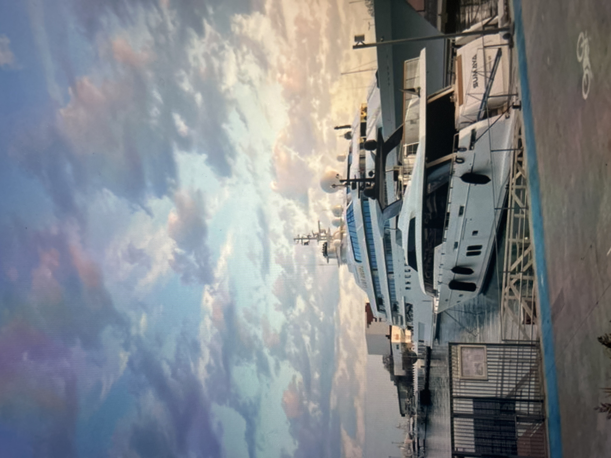 The image, appearing rotated 90 degrees counter-clockwise, showcases a large white yacht docked in a marina under a dramatic sky. The scene is illuminated by the warm, soft light characteristic of either sunrise or sunset, casting hues of blue, purple, pink, and orange across the voluminous clouds.
The primary subject is a multi-deck superyacht, predominantly white with dark windows and a sleek design. It is equipped with several radar domes and communication antennae on its upper levels. The yacht is moored alongside a dock or pier, with calm blue water visible between the vessel and the dock's edge. In the foreground, the dock features dark metal railings and a gate. A white bicycle symbol is painted on the paved surface of the dock, suggesting a designated path.
Near the stern of the main yacht, a smaller boat or tender is covered with a white canvas, prominently displaying the name "SUMAYA." Indistinct text can also be observed on the hull of the main yacht near its bow. The background reveals additional boats and harbor structures.
There are no people visible, allowing the focus to remain on the luxurious yacht and the striking, colorful sky. The overall impression is one of tranquility and grandeur in a well-appointed port setting.