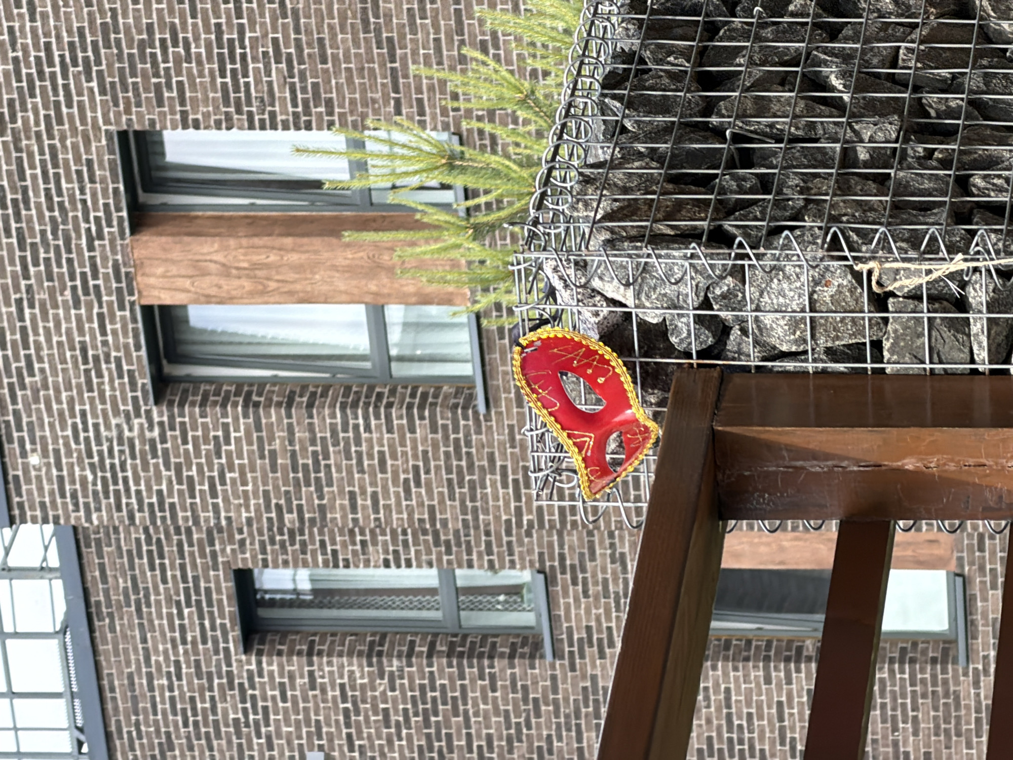 An outdoor scene in Strelna, Russia, features a brick building with multiple windows. In the foreground, a wooden structure is visible, supporting a gabion wall filled with stones. A red and gold masquerade mask is attached to the wire mesh of the gabion wall. Several small green evergreen saplings grow behind the wall.