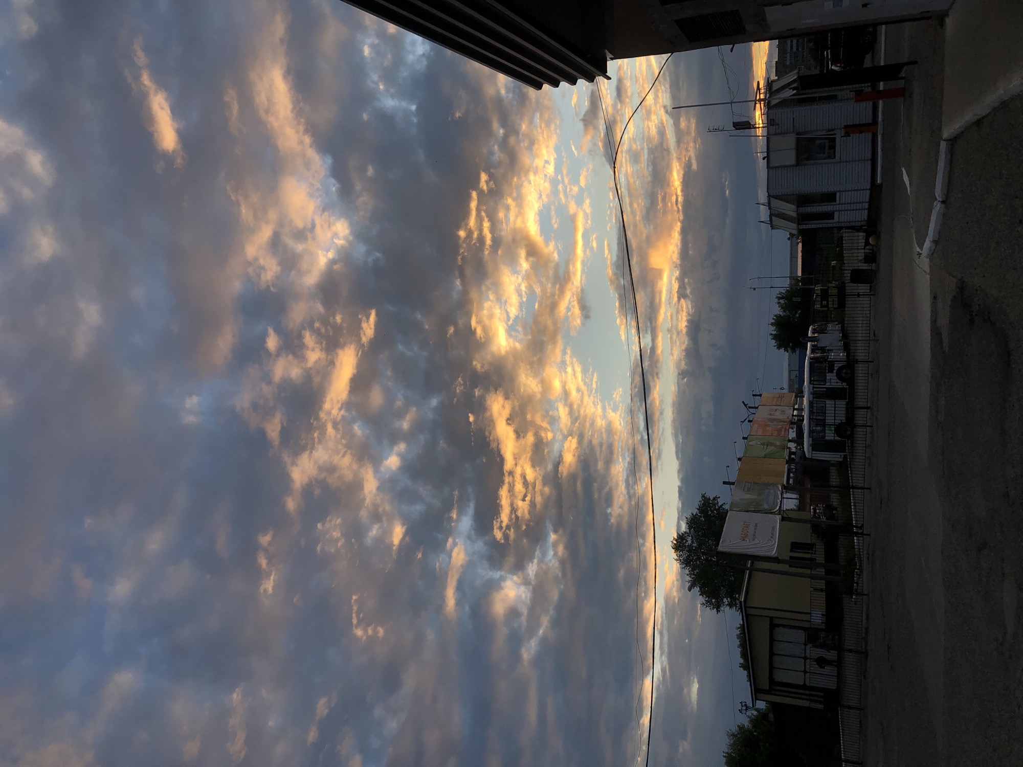 The image shows an exterior view of a bus depot or similar facility in the city of Chaikovsky, Russia, during a sunset. The sky is filled with dramatic clouds, illuminated with hues of orange and yellow from the setting sun, contrasting with darker grey and blue clouds. In the foreground, there is a paved area with some concrete curbs. To the left, a yellow building with a large garage-style door is visible, with a black metal fence in front of it. A white bus is parked to the right of this building, behind the fence. Further to the right, there is a small white booth-like structure, possibly a security checkpoint, with an orange bollard next to it. In the distance, more buildings and trees are faintly visible, and a power line stretches across the frame. The overall scene depicts a somewhat industrial or service area bathed in the warm light of the sunset.