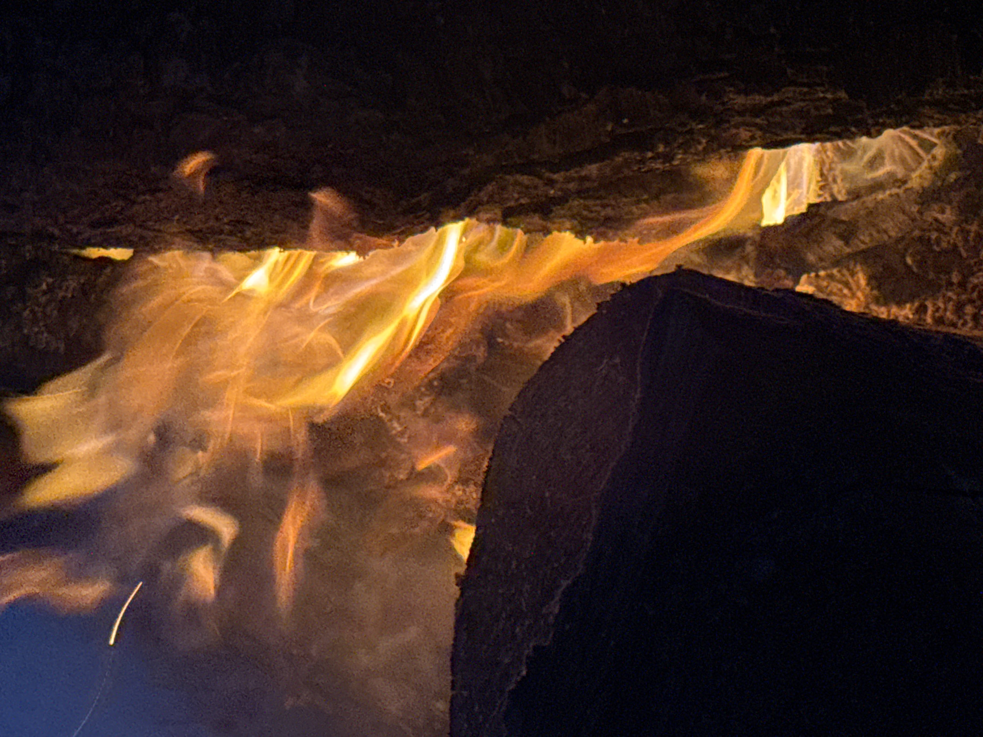 The image displays a close-up, low-angle perspective of a bonfire or campfire at night. Dominant bright orange and yellow flames surge upwards from between two large, dark logs. On the left, a large, dark, roughly triangular log with a rounded upper edge occupies the lower left quadrant, partially obscuring the base of the fire. Its surface shows subtle textural variations. On the right, a dark, vertical, textured log or piece of wood forms a boundary for the flames. The intense, warm light from the fire illuminates the immediate surroundings, casting an orange glow on the inner surfaces of the logs and the smoke. The top left corner reveals a dark blue or deep indigo hue, possibly indicating the night sky, with a single bright, horizontal streak of light, likely a glowing ember or spark in motion. The overall environment is dark, emphasizing the intense luminosity of the fire. There are no individuals or other identifiable objects present in the scene.