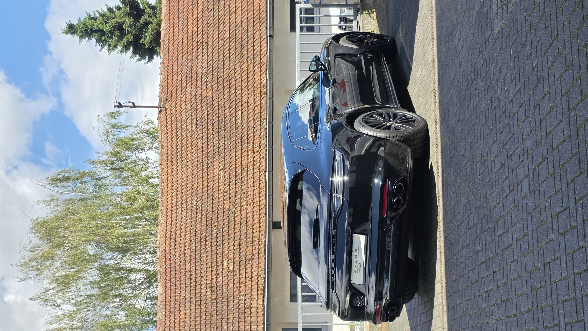A black Porsche Panamera Sport Turismo is parked on a brick driveway in front of a building with a red tile roof. The car is viewed from the rear, angled slightly to the left. The building appears to be a house or a small commercial establishment. Trees are visible in the background, along with a blue sky with scattered clouds. A metal fence runs along the front of the building. The car has a "Porsche Approved" sticker on the rear bumper.