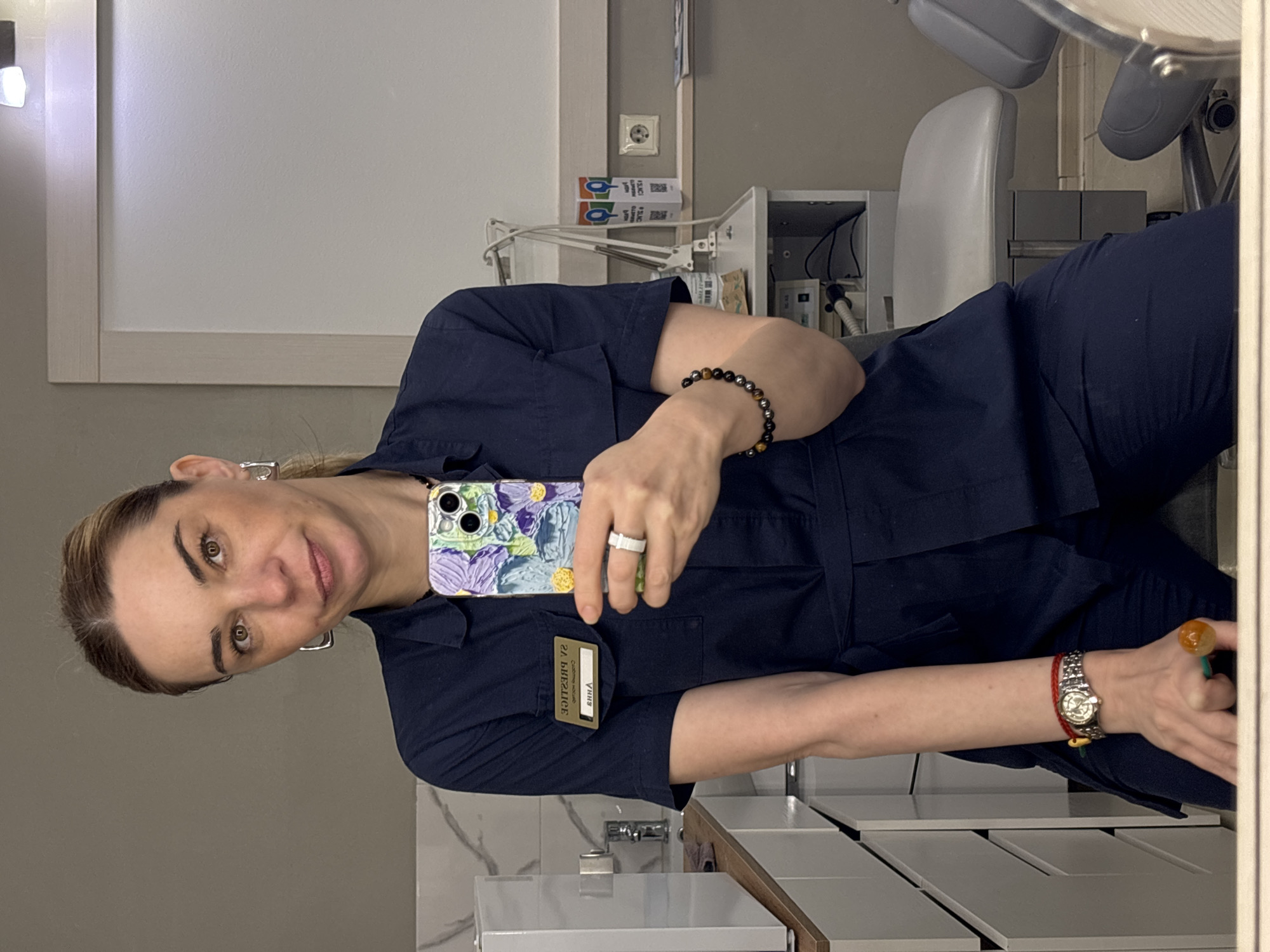 A woman is taking a selfie in a room that appears to be part of a dental office or clinic. She is wearing a navy blue uniform with a name tag labeled "ERINA V." (The name is partially obscured due to its orientation). She holds a phone with a patterned case. A bracelet is on her wrist. A pen or small object is in her hand. The background includes a dental chair, cabinets, and other equipment associated with dental care. The image location is identified as Vsevolozhsk, Russia, based on the identification data.