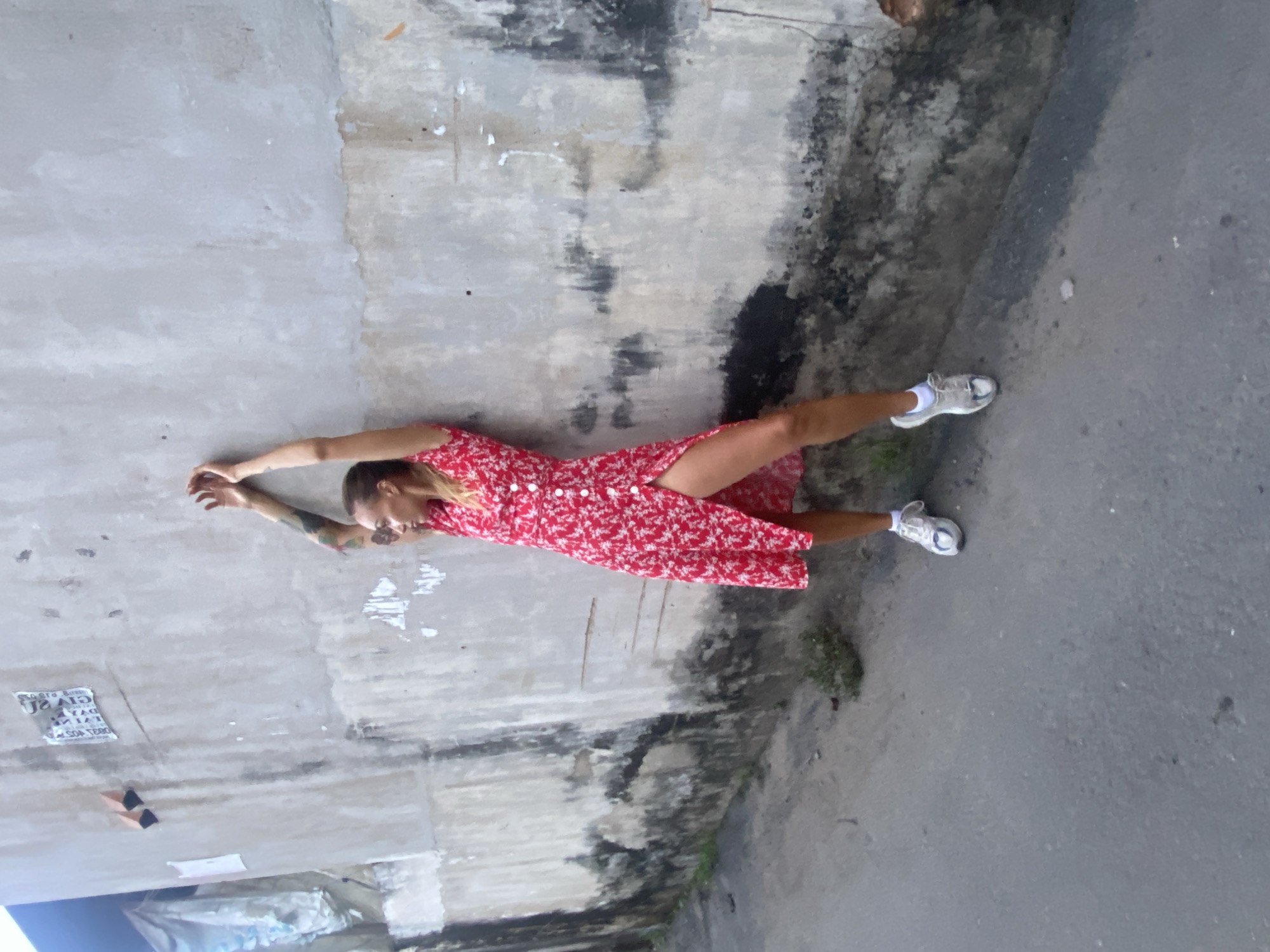A woman lies against a concrete wall next to a street in Đồng Nai, Vietnam. She is wearing a red floral dress and white sneakers. Her arms are raised above her head. The wall is weathered and stained. The street surface is gray and appears to be concrete or asphalt.